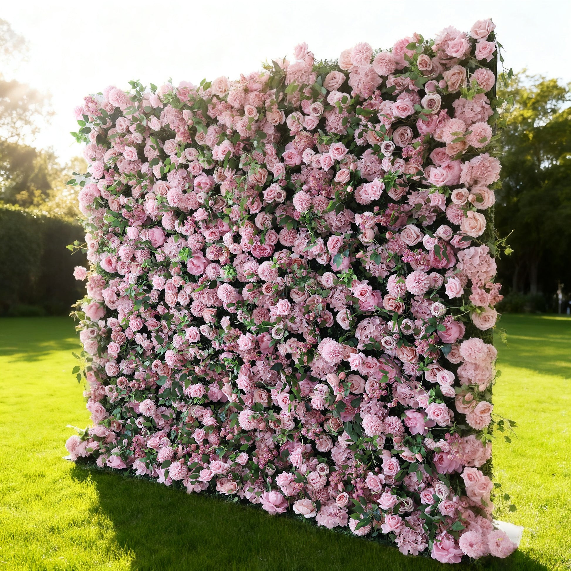 This image features a large, square floral wall outdoors on a lush green lawn. It's densely covered with an array of pink flowers, creating a rich, romantic display. Bathed in sunlight, with trees and a clear sky in the background, the scene exudes a sense of natural beauty, elegance, and charm, perfect for decorative purposes in events or as a picturesque backdrop.