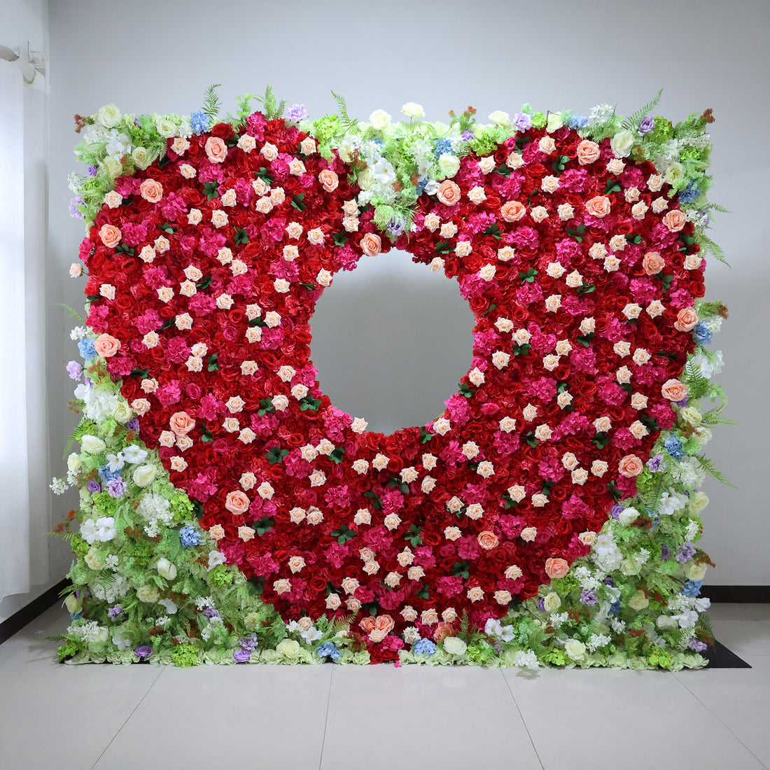 This vibrant indoor floral wall from **Valar Flowers** is a lush tapestry of deep red, blush pink, and creamy white blooms—including roses and peonies—interwoven with green foliage and subtle blue/purple accents. A striking circular cutout at its center creates a dramatic focal point, evoking a bold, romantic, and sophisticated atmosphere ideal for weddings, photo backdrops, or elegant celebrations.