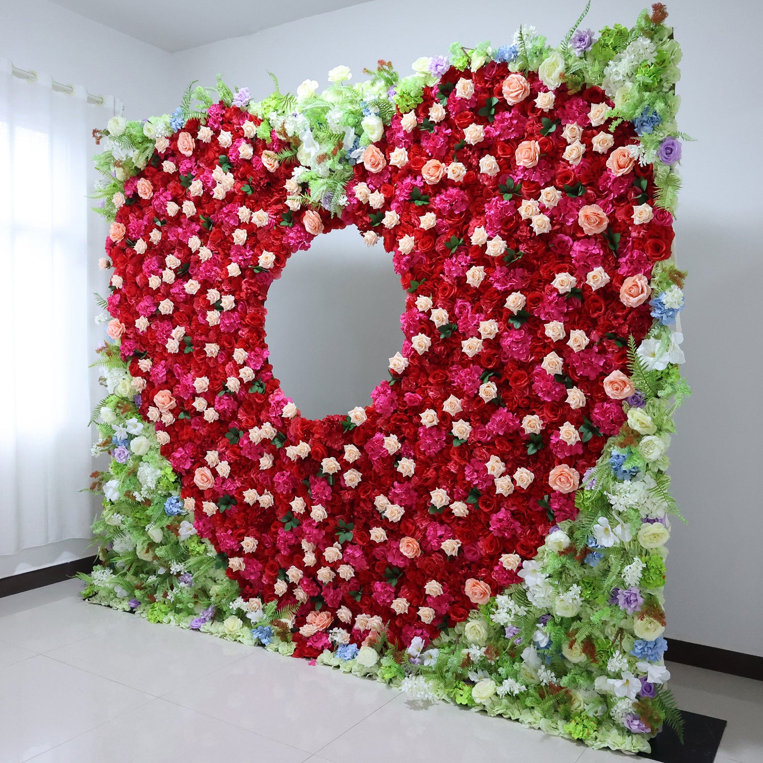 This vibrant indoor floral wall from **Valar Flowers** is a lush, eye-catching tapestry of deep red, blush pink, and creamy white blooms—including roses, peonies, and delicate filler flowers—interwoven with vibrant green foliage and subtle blue and purple accents. The design features a striking circular cutout at its center, creating a dramatic focal point. 