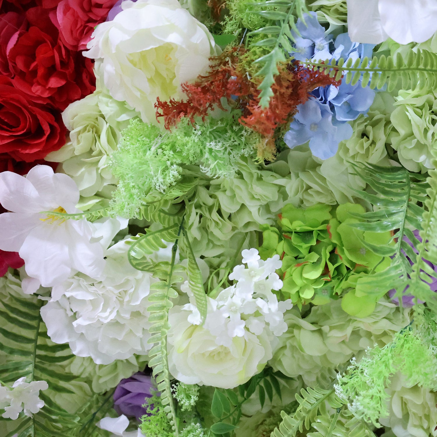 This close-up from Valar Flowers features a lush, multi-textured floral arrangement with a vibrant palette. It includes deep red roses, creamy white peonies and roses, soft green hydrangeas, delicate blue hydrangeas, and feathery green ferns, accented by subtle reddish-brown foliage. The dense, layered design creates a rich, three-dimensional texture, evoking a fresh, romantic, and naturally elegant atmosphere ideal for weddings, photo backdrops, or elegant celebrations.
