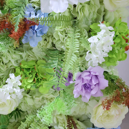 This close-up from Valar Flowers showcases a lush, textured floral arrangement with a fresh, pastel palette. It features soft green hydrangeas, delicate blue hydrangeas, a rich purple rose, and vibrant white lilac-like blooms, all interwoven with feathery green ferns and subtle reddish-brown accents. The dense, layered design creates a rich, three-dimensional texture, evoking a fresh, romantic, and naturally elegant atmosphere ideal for weddings, photo backdrops, or elegant celebrations.