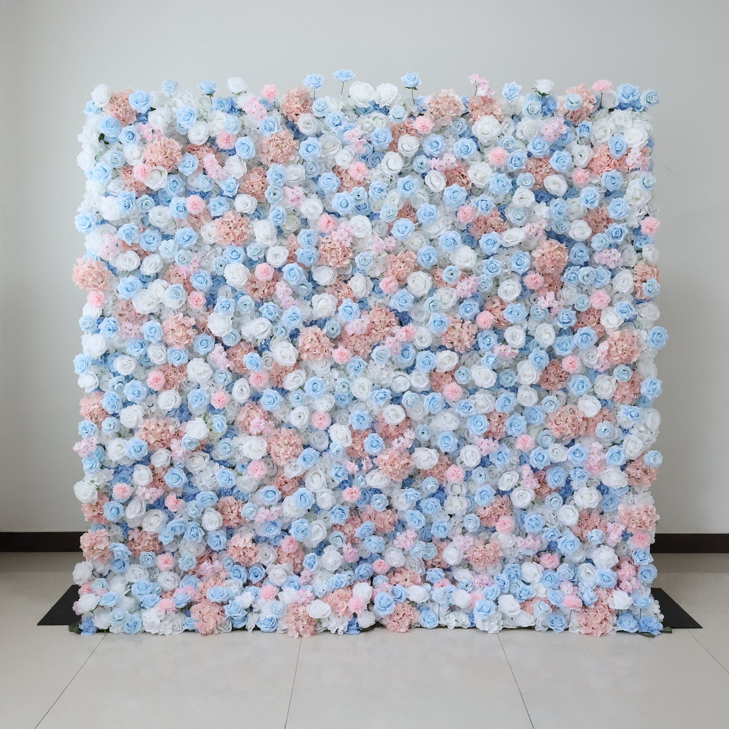 This image showcases a large, square floral wall indoors. It's densely covered with an array of artificial flowers in soft pink, light blue, and white hues. The vibrant blooms create a rich, colorful, and romantic display. Set against a plain wall with a tiled floor, the scene exudes a sense of beauty, elegance, and charm, perfect for decorative purposes in events or as a striking interior backdrop.