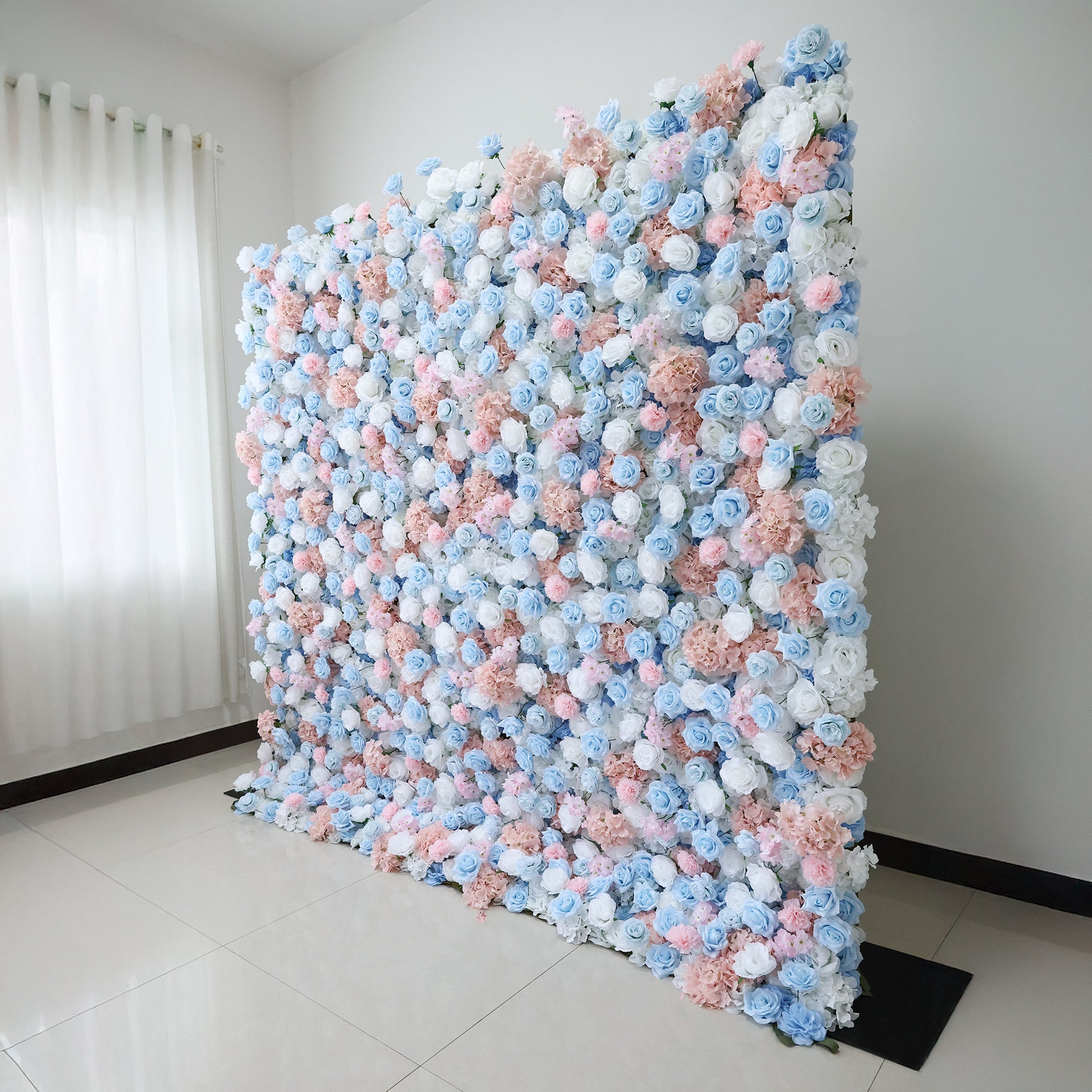 This image features a large, artificial floral wall indoors. It's densely covered with an array of flowers in soft pink, light blue, and white hues. The vibrant blooms create a rich, colorful, and romantic display. Set in a room with white walls, a tiled floor, and a curtain allowing light in, the scene exudes a sense of beauty, elegance, and charm, perfect for decorative purposes in events or as a striking interior backdrop.