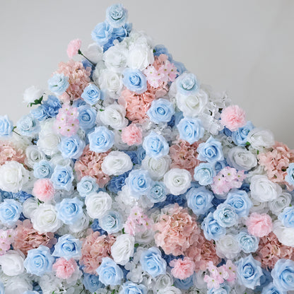 This image showcases a dense arrangement of artificial flowers in soft hues of blue, white, and pink. The blooms, including roses and other floral varieties, are clustered together to form a lush, romantic display. Set against a plain background, the pastel colors and intricate petals create an elegant and charming scene, perfect for decorative purposes in events or as a visually appealing artistic piece.