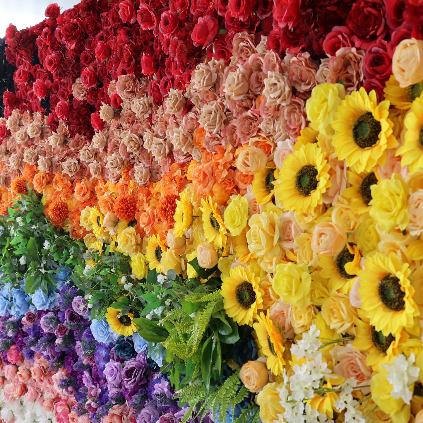 This vibrant, close-up view of a floral wall features a bold, sunset-to-rainbow gradient. It transitions from deep red and blush peach blooms at the top, through sunny yellow sunflowers and lush green foliage, to cool blue and purple flowers, and soft pink and white roses at the bottom.
The dense, textured arrangement of artificial flowers exudes joyful, vibrant energy, making it a lively focal point for pride events, festivals, or colorful celebrations.
