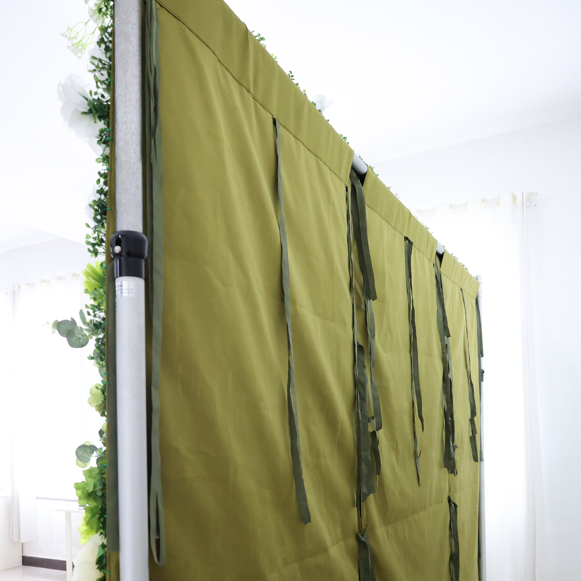 This image shows the side of a structure, likely part of a floral or decorative display setup. It features an olive - green fabric panel attached to a metal frame. Several green straps hang from the fabric, possibly for securing or adjusting the panel. On the left edge, there are artificial green leaves and white flowers, adding a touch of nature. The setting appears to be indoors, with bright lighting and a white - walled background, giving a clean and organized look, suggesting preparation for an event or
