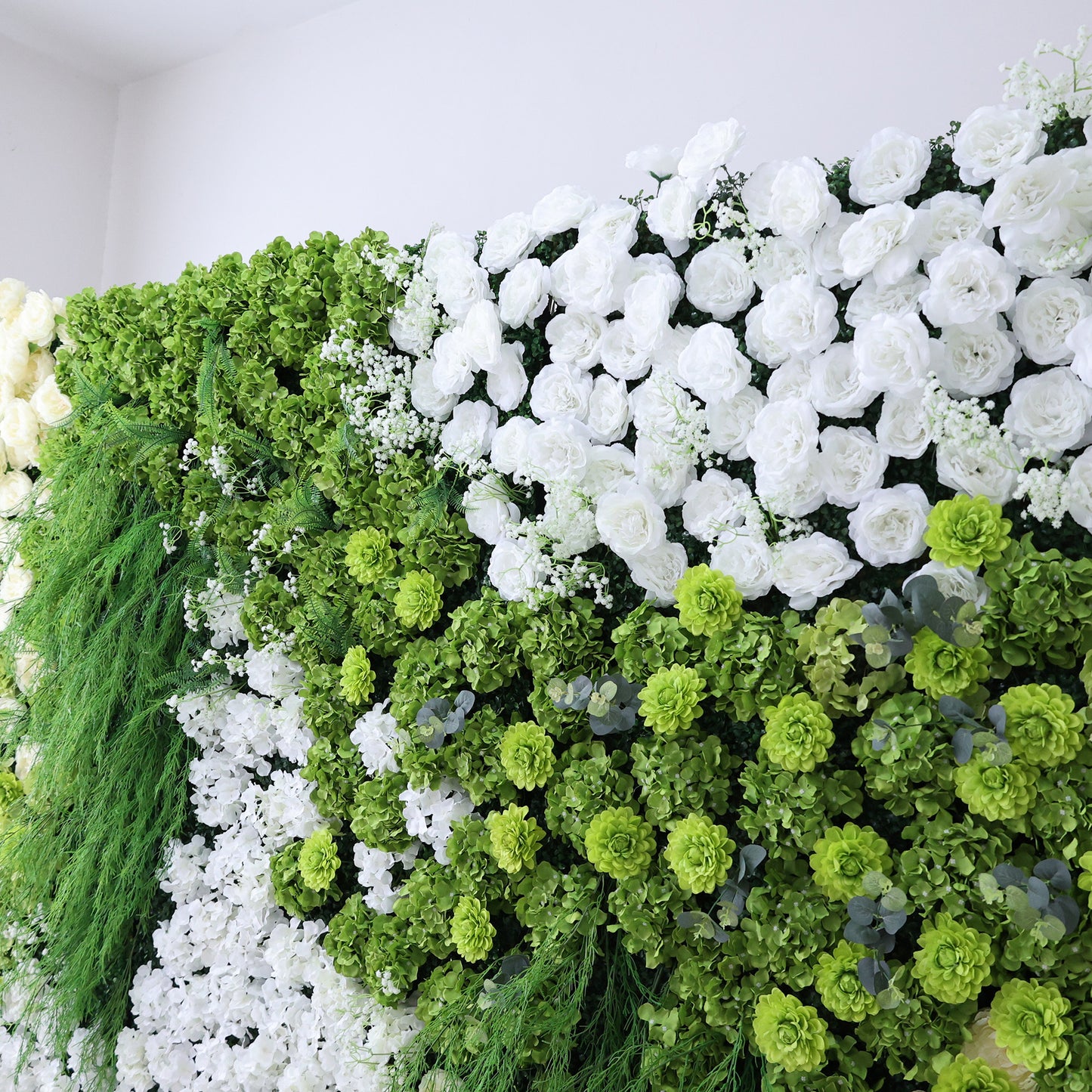This image features a lush, artificial floral wall indoors. It combines various white flowers with abundant green foliage and textured green blooms. The dense arrangement creates a vibrant, natural - looking display. Set against a plain white wall, the scene exudes a sense of freshness, harmony, and elegance, perfect for decorative purposes in events or as a striking interior backdrop, blending artistry with a touch of nature's charm.