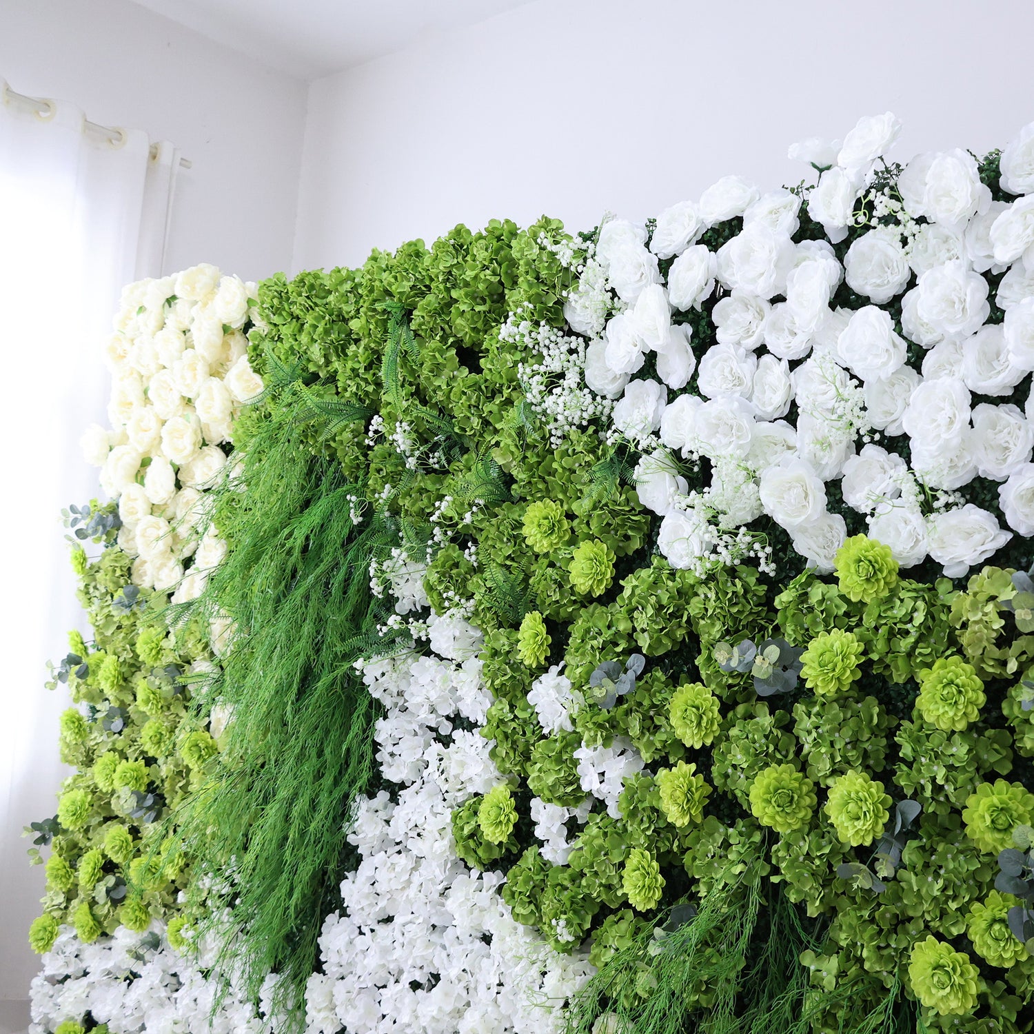 This image features a lush, artificial floral wall indoors. It combines various white flowers with abundant green foliage and textured green blooms. The dense arrangement, including trailing greenery and small white accents, creates a vibrant, natural - looking display. Set against a plain white wall with light filtering through a curtain, the scene exudes freshness, harmony, and elegance, perfect for event decor or as a striking interior backdrop, blending artistry with nature&