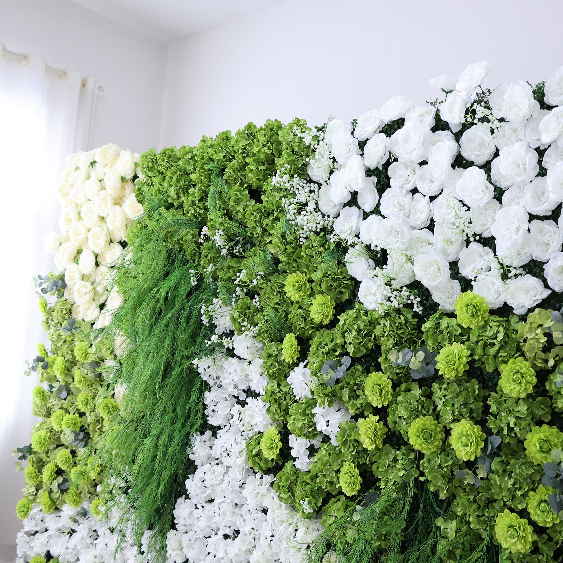 This image features a lush, artificial floral wall indoors. It combines various white flowers with abundant green foliage and textured green blooms. The dense arrangement, including trailing greenery and small white accents, creates a vibrant, natural - looking display. Set against a plain white wall with light filtering through a curtain, the scene exudes freshness, harmony, and elegance, perfect for event decor or as a striking interior backdrop, blending artistry with nature's charm.
