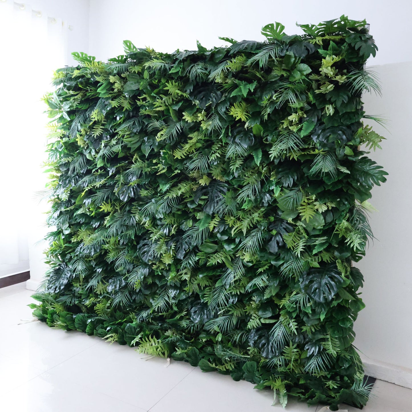 This Valar Flowers "flower wall" blends varied foliage—deep green monstera leaves, delicate ferns, and tropical fronds—in layered, textured shades of green. Set indoors against plain white walls and light flooring, soft ambient light highlights its dense, organic depth. Though bloom-free, it embodies Valar’s nature-focused styling: ideal as an indoor event backdrop, balancing fresh, earthy texture with versatile, elegant decor for modern or tropical themes.
