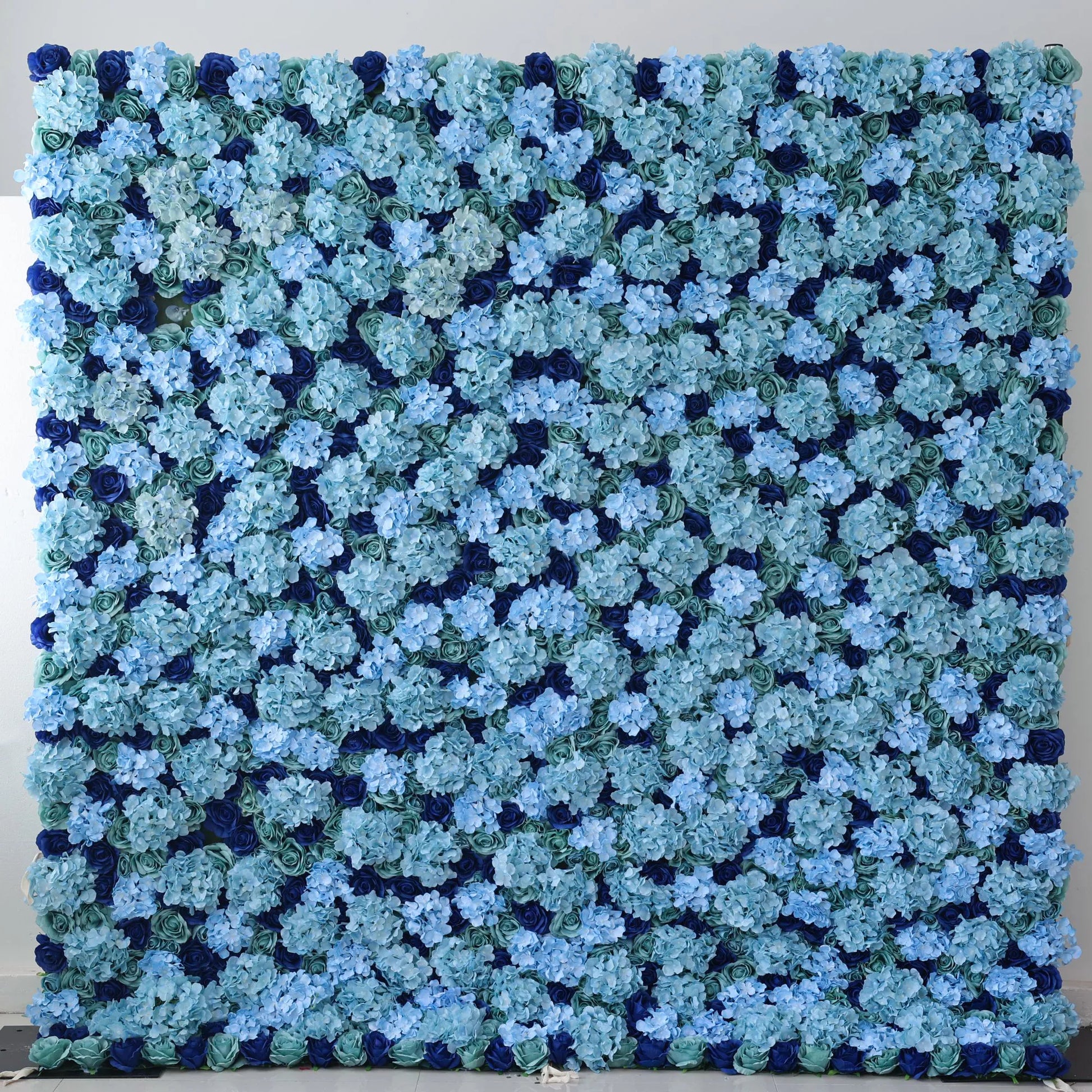 This large artificial flower wall features a lush arrangement of blue hydrangeas in varying shades, layered over green roses and dark blue blossoms. The dense, harmonious color blend creates an elegant, serene backdrop, perfect for adding a refined, decorative touch to events or spaces. 
