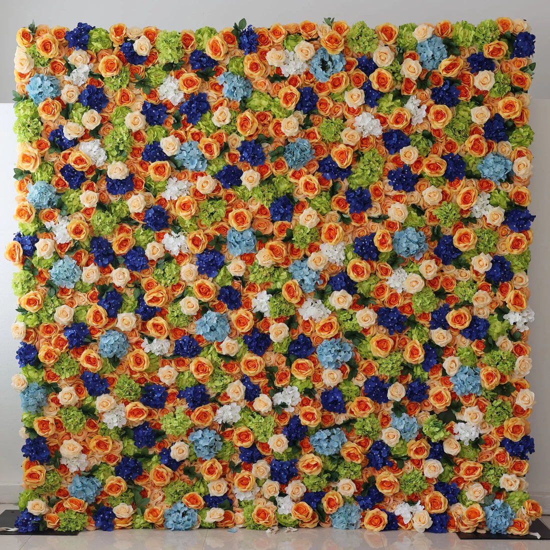 This large artificial flower wall bursts with vibrant colors. Orange roses, blue hydrangeas, white blooms, and green foliage are densely arranged, creating a lush, textured display. The harmonious color mix brings a cheerful, lively vibe, perfect as a backdrop for weddings, parties, or photo shoots, adding an eye - catching, decorative charm.