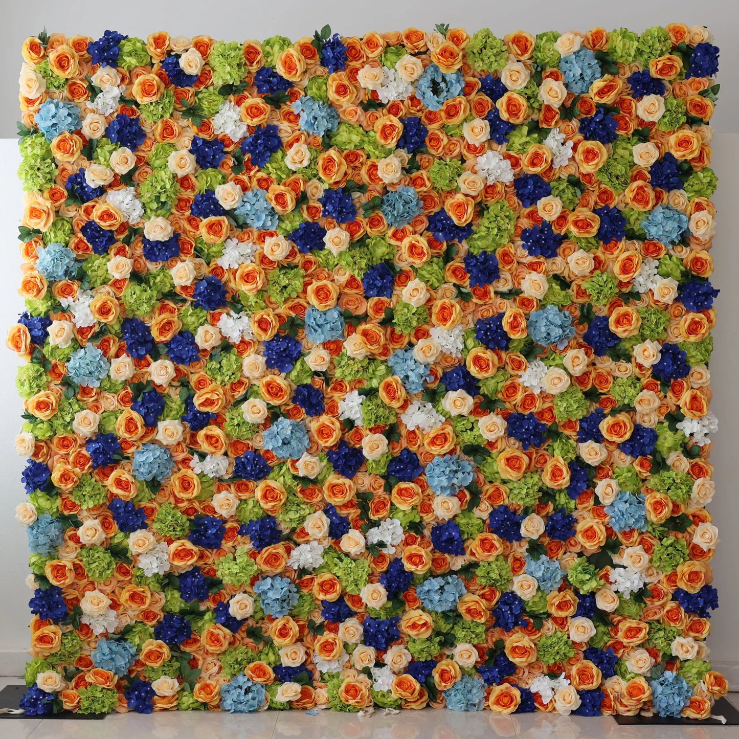This large artificial flower wall bursts with vibrant colors. Orange roses, blue hydrangeas, white blooms, and green foliage are densely arranged, creating a lush, textured display. The harmonious color mix brings a cheerful, lively vibe, perfect as a backdrop for weddings, parties, or photo shoots, adding an eye - catching, decorative charm. 
