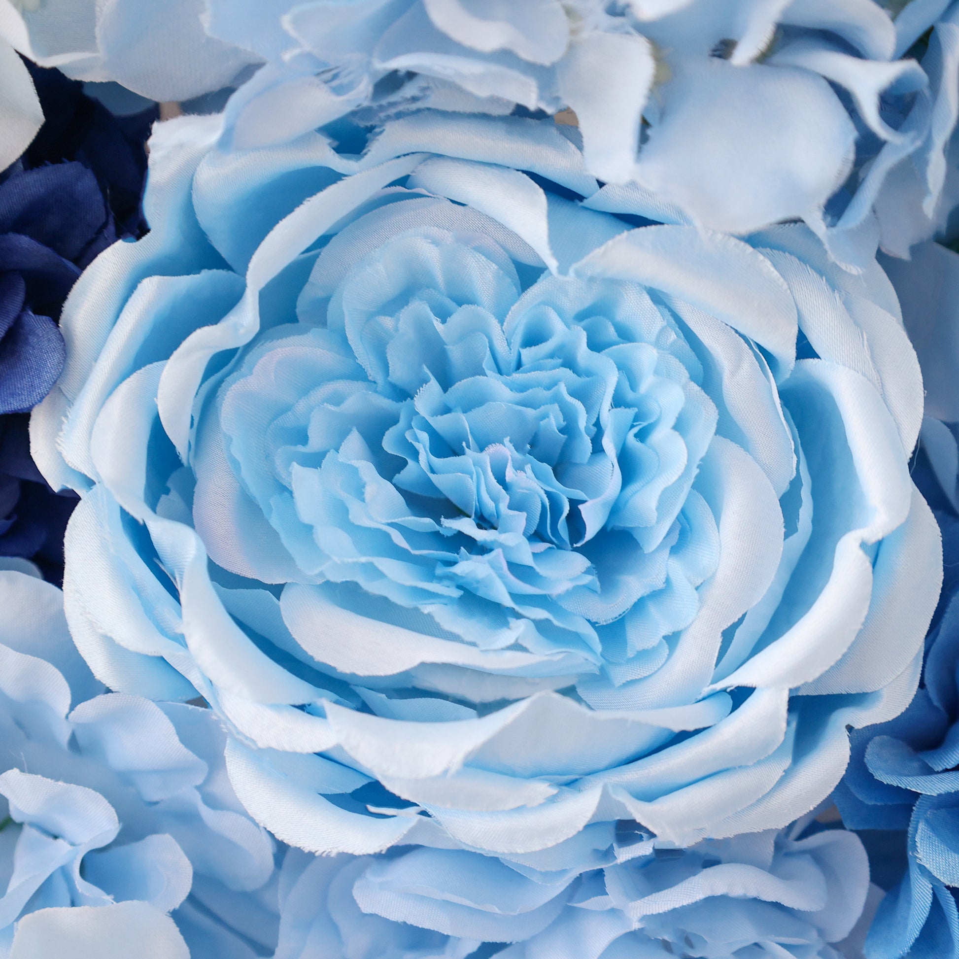 A close - up of a large artificial blue rose with layered, soft - toned petals. Surrounding it are other light blue and dark blue blooms, creating a lush, elegant floral arrangement. The intricate petal details and serene color palette evoke a calm, sophisticated beauty. 