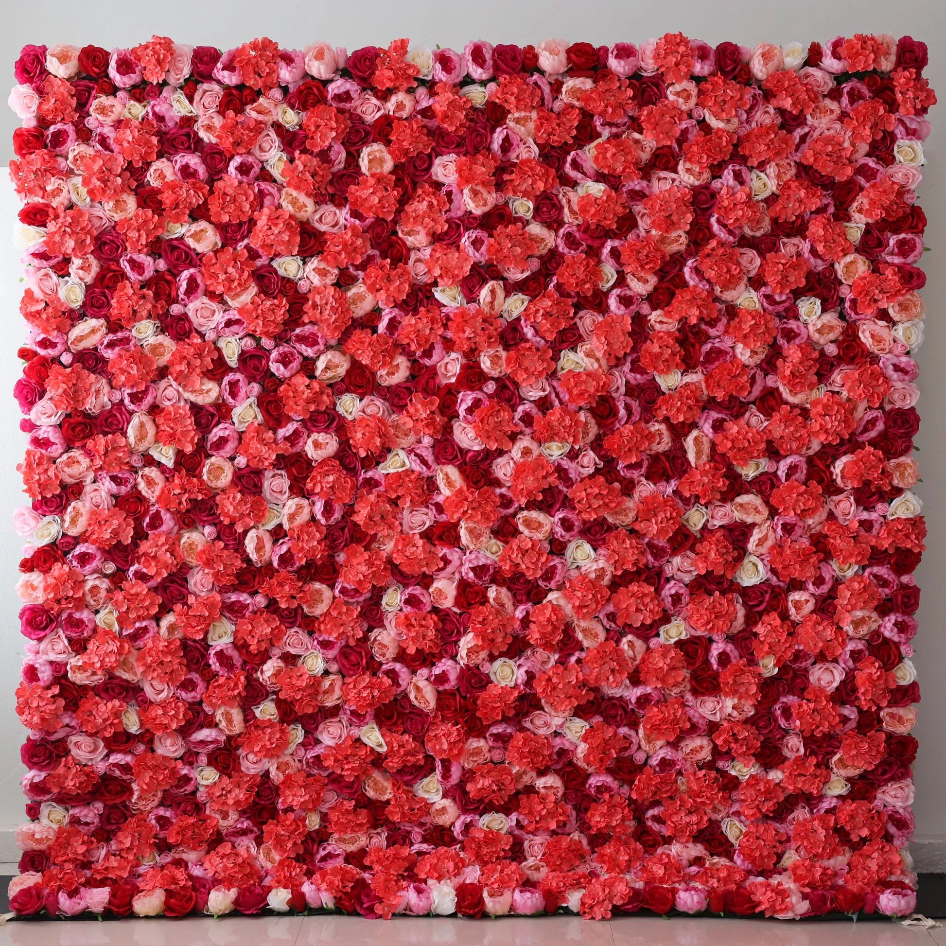 A large artificial flower wall showcases a vibrant mix of roses in shades of coral, pink, white, and deep red. The dense, layered arrangement creates a lush, romantic texture, with blooms tightly packed to form a striking, colorful backdrop. Perfect for adding a lively, decorative touch to events or spaces, exuding warmth and elegance. 