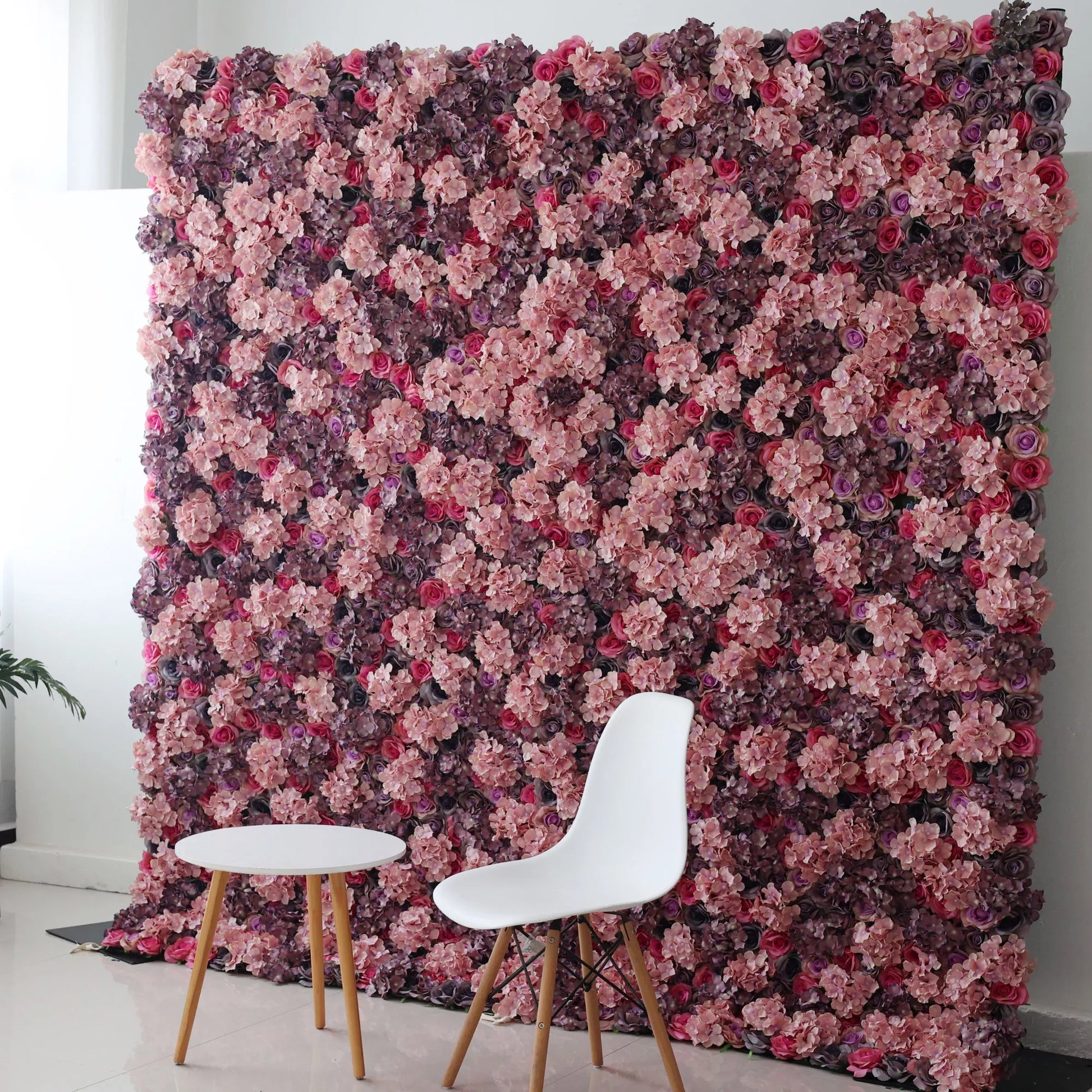 Valar Flowers Roll Up Backdrop: This picture shows a large artificial flower wall in rich hues of purple, pink, and red, with various blooms densely arranged. In front, a minimalist white chair and a small round table with wooden legs create a contrast. The setup blends natural - inspired decor with modern simplicity, ideal for photo backdrops or stylish event spaces, offering an elegant and romantic atmosphere. 