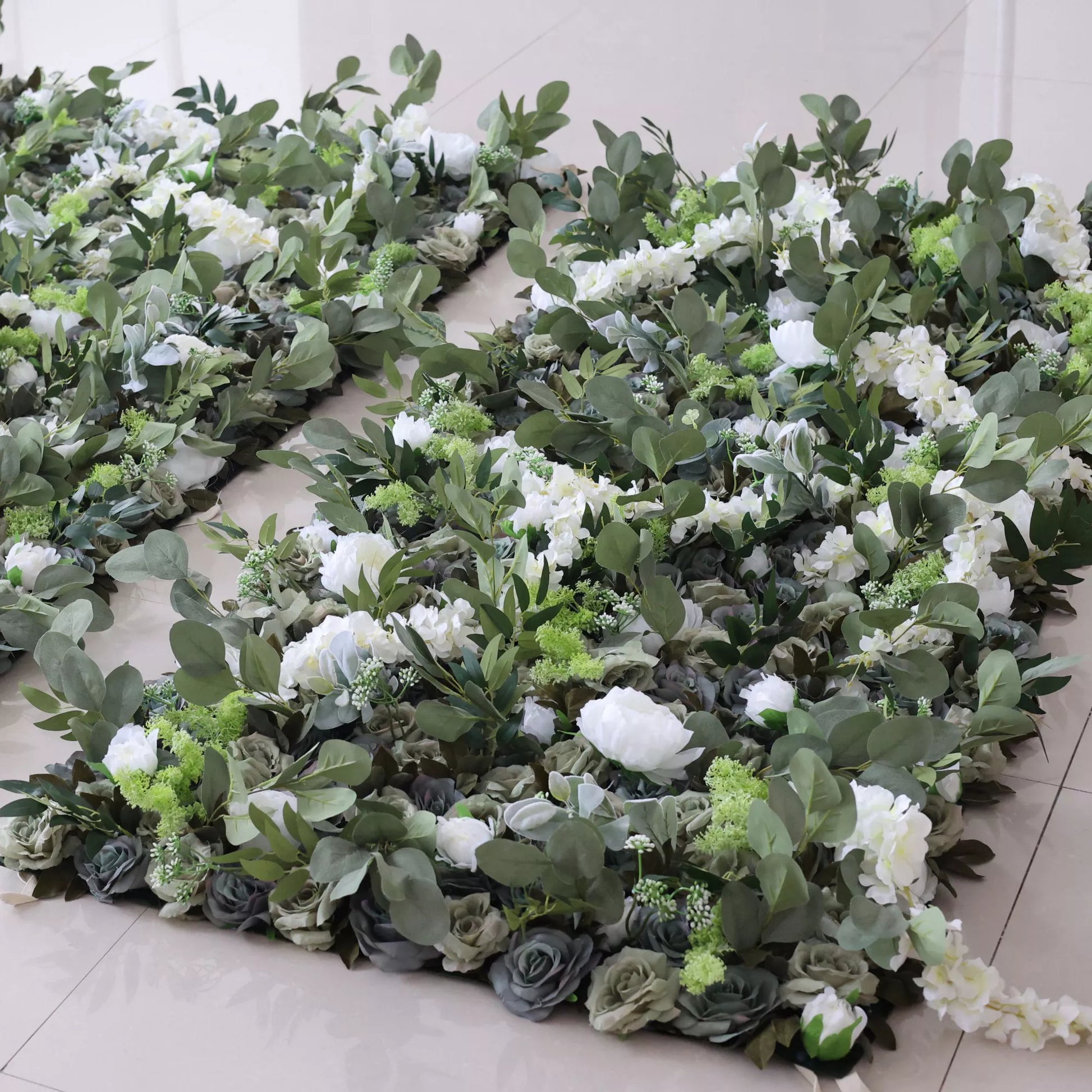 This picture shows several pieces of artificial floral decor. They feature a mix of white blooms, green foliage, and gray roses, arranged on a light - colored tiled floor. A small blue circle is visible in the center, adding a distinct detail. The lush, layered design creates an elegant, natural - looking display, perfect for decorative use. 