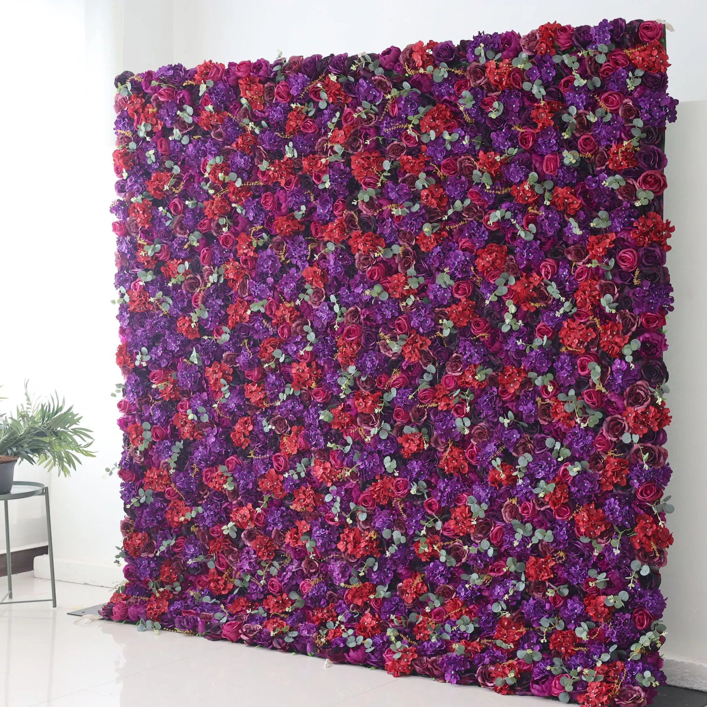 This picture features a large artificial flower wall, a stunning display of deep purple and vibrant red blooms. Interspersed with small green leaves, the dense arrangement creates a rich, luxurious texture. The contrasting colors and lush design make it an eye - catching backdrop, perfect for events like weddings or parties, adding a touch of elegance and drama to the space. A small plant on the side complements the floral scene. 