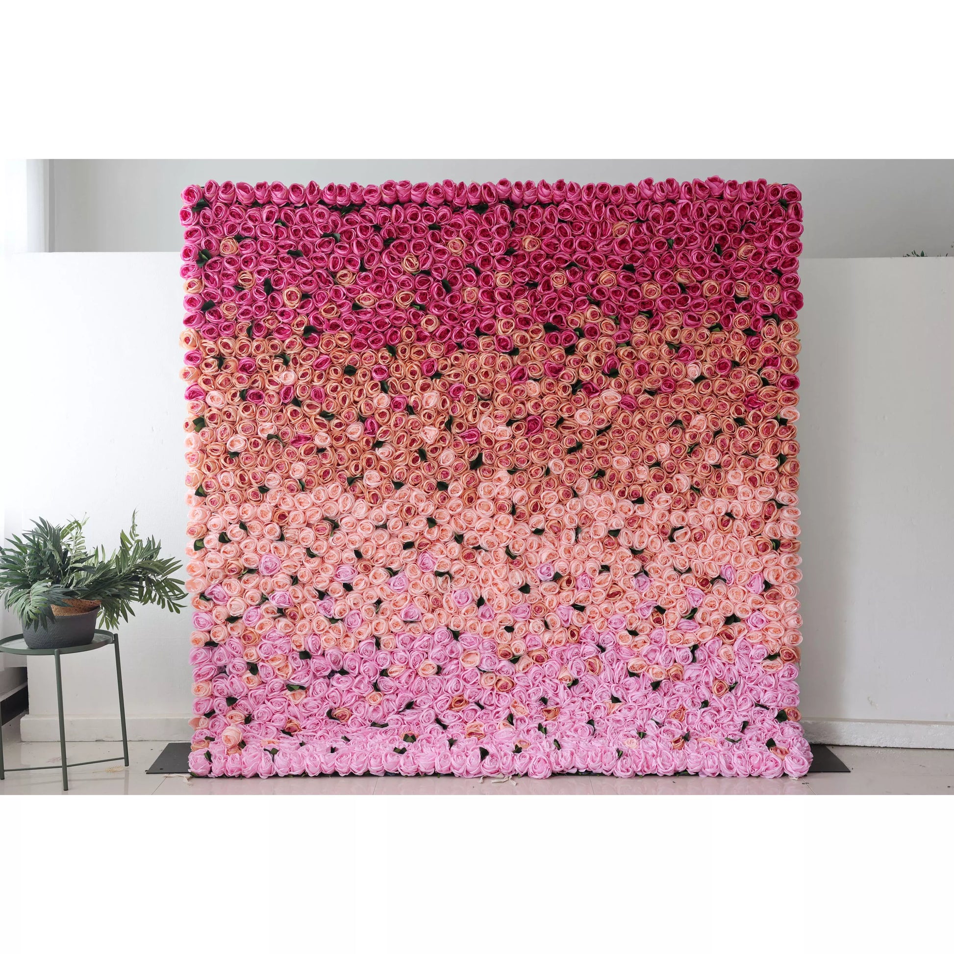 This picture shows a large artificial flower wall, a beautiful gradient of pink shades. It’s densely packed with rose - like blooms, transitioning from deep pink at the top to soft pink at the bottom. The uniform, lush arrangement creates a romantic, eye - catching display, perfect for event backdrops. A small green plant on the side adds a touch of natural contrast, enhancing the overall decorative appeal of the space. 