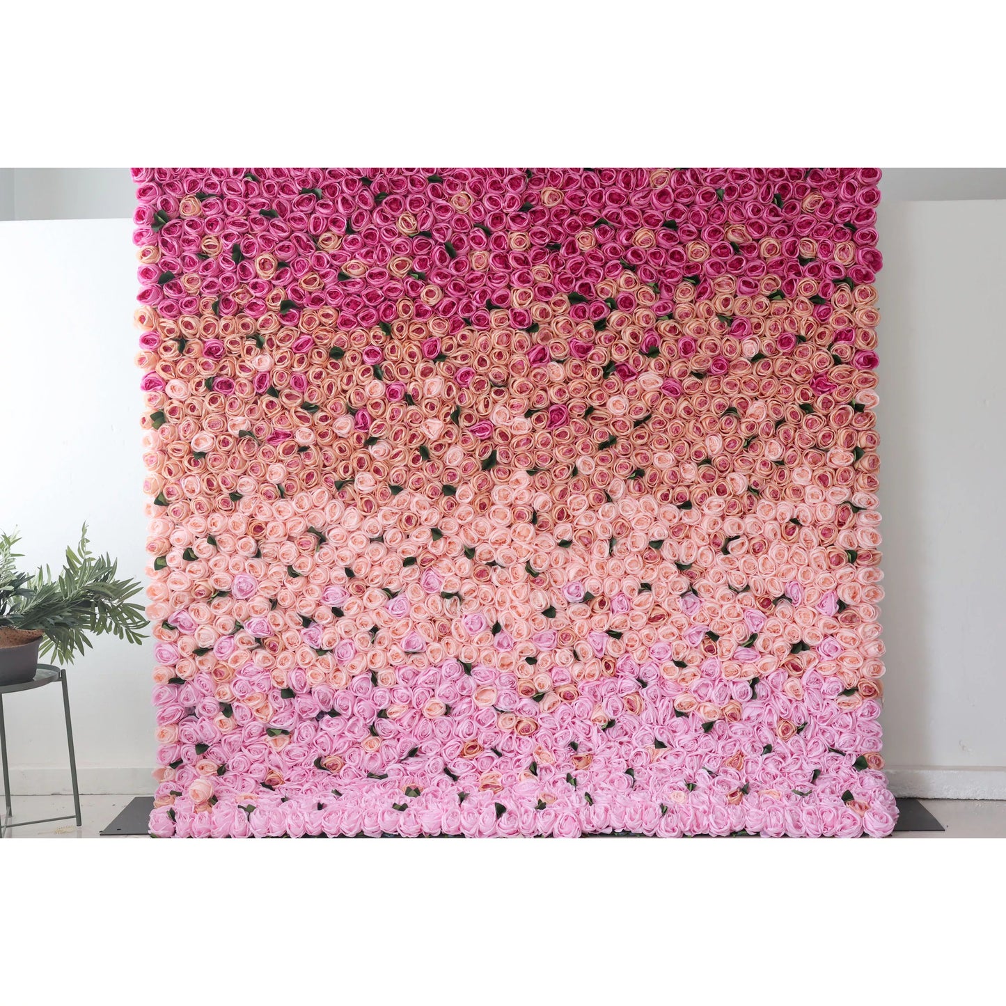 This picture showcases a large artificial flower wall, a beautiful gradient of pink tones. It’s densely packed with rose - like blooms, transitioning from deep pink at the top to soft pink at the bottom. The uniform, lush arrangement creates a romantic, eye - catching display, perfect for event backdrops. A small green plant on the side adds a touch of natural contrast, enhancing the overall decorative appeal of the space. 