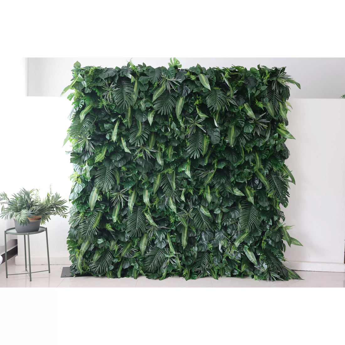 This picture presents a large, lush artificial greenery wall. It features a dense arrangement of diverse green leaves, including broad palm - like fronds and smaller, variegated foliage. The rich, verdant texture creates a natural, tropical - inspired backdrop. Placed against a plain white wall and floor, with a small plant visible on the side, it brings an instant touch of nature indoors, ideal for decorative uses in events or interior spaces.