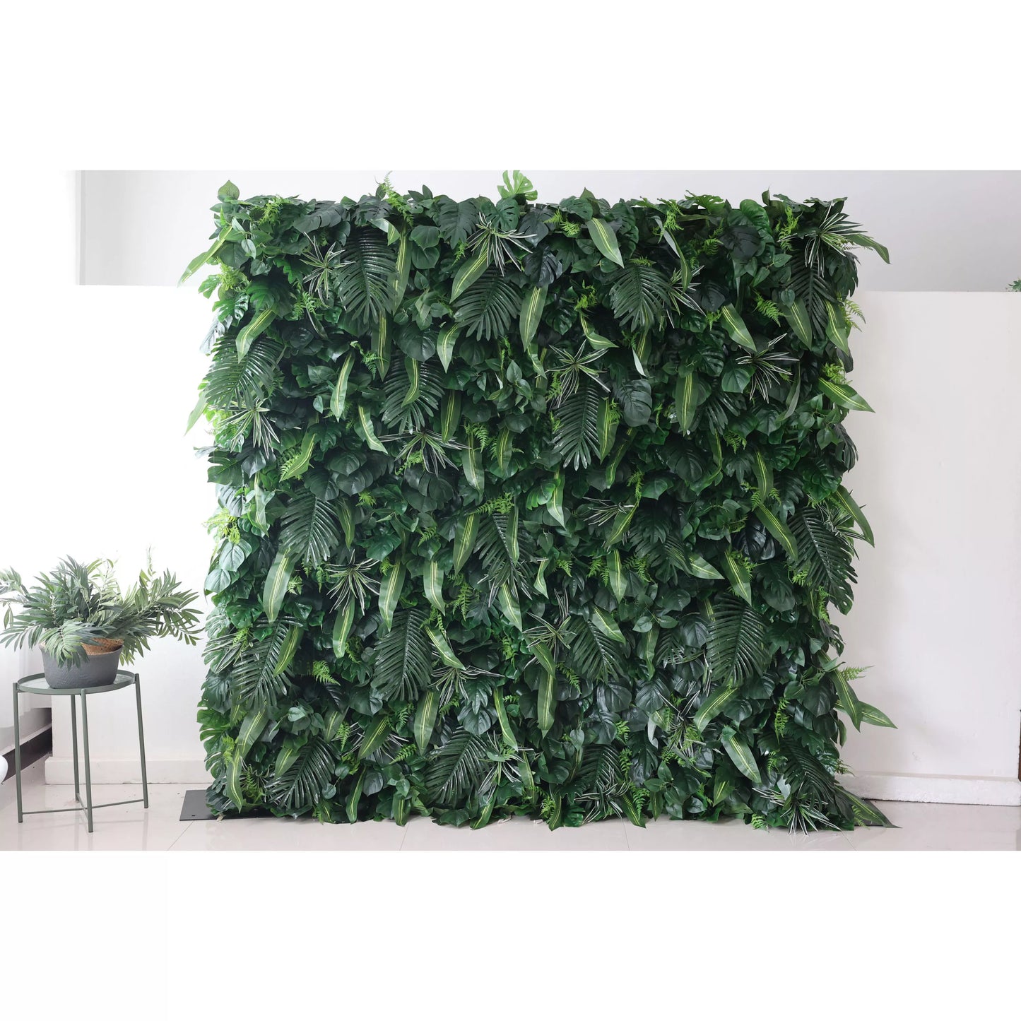 This picture presents a large, lush artificial greenery wall. It features a dense arrangement of diverse green leaves, including broad palm - like fronds and smaller, variegated foliage. The rich, verdant texture creates a natural, tropical - inspired backdrop. Placed against a plain white wall and floor, with a small plant visible on the side, it brings an instant touch of nature indoors, ideal for decorative uses in events or interior spaces. 
