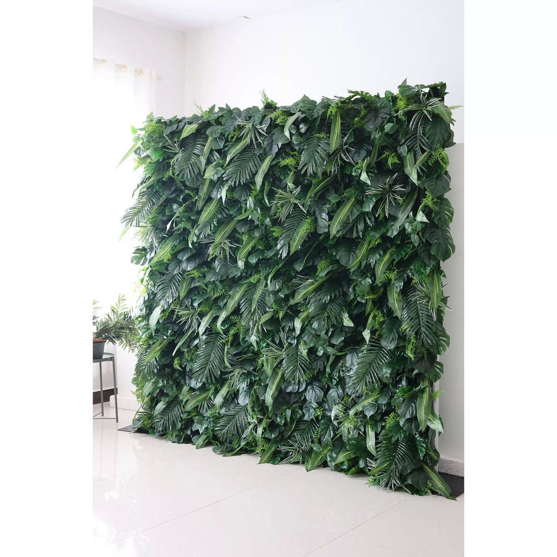 This picture showcases a large, lush artificial greenery wall. It features a dense arrangement of various green leaves, including ferns and broad - bladed plants. The rich, verdant foliage creates a natural, tropical - like backdrop. Placed against a white wall and floor, with a small plant on a side table, it adds a touch of nature to the indoor space, ideal for decorative purposes in events or interiors.