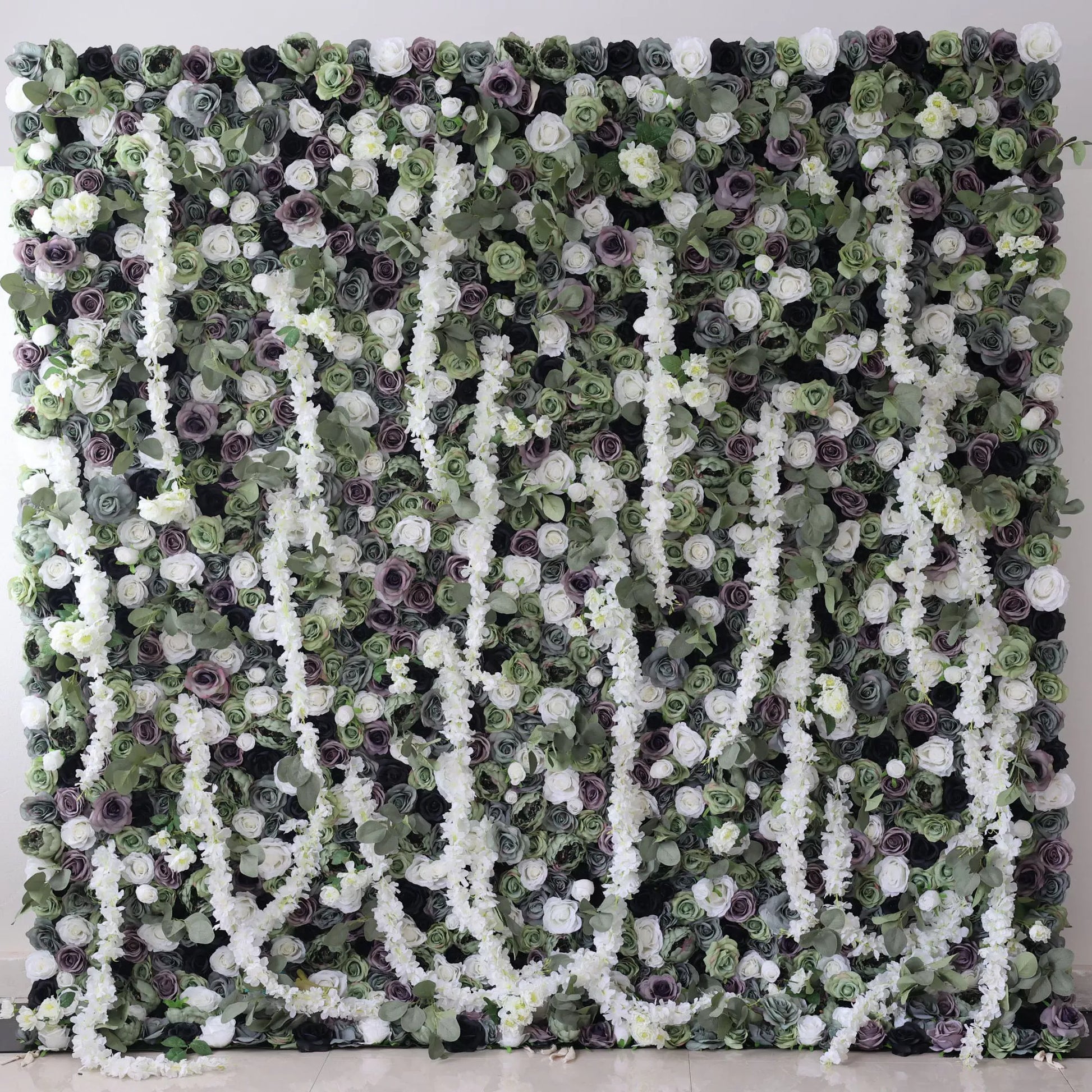This is a large artificial flower wall with a green - dominated color scheme. It features white blossoms, purple roses, and dense green foliage. The arrangement includes cascading white floral vines, creating a lush, natural - looking display. Ideal for decorative event backdrops, adding an elegant and verdant touch. 
