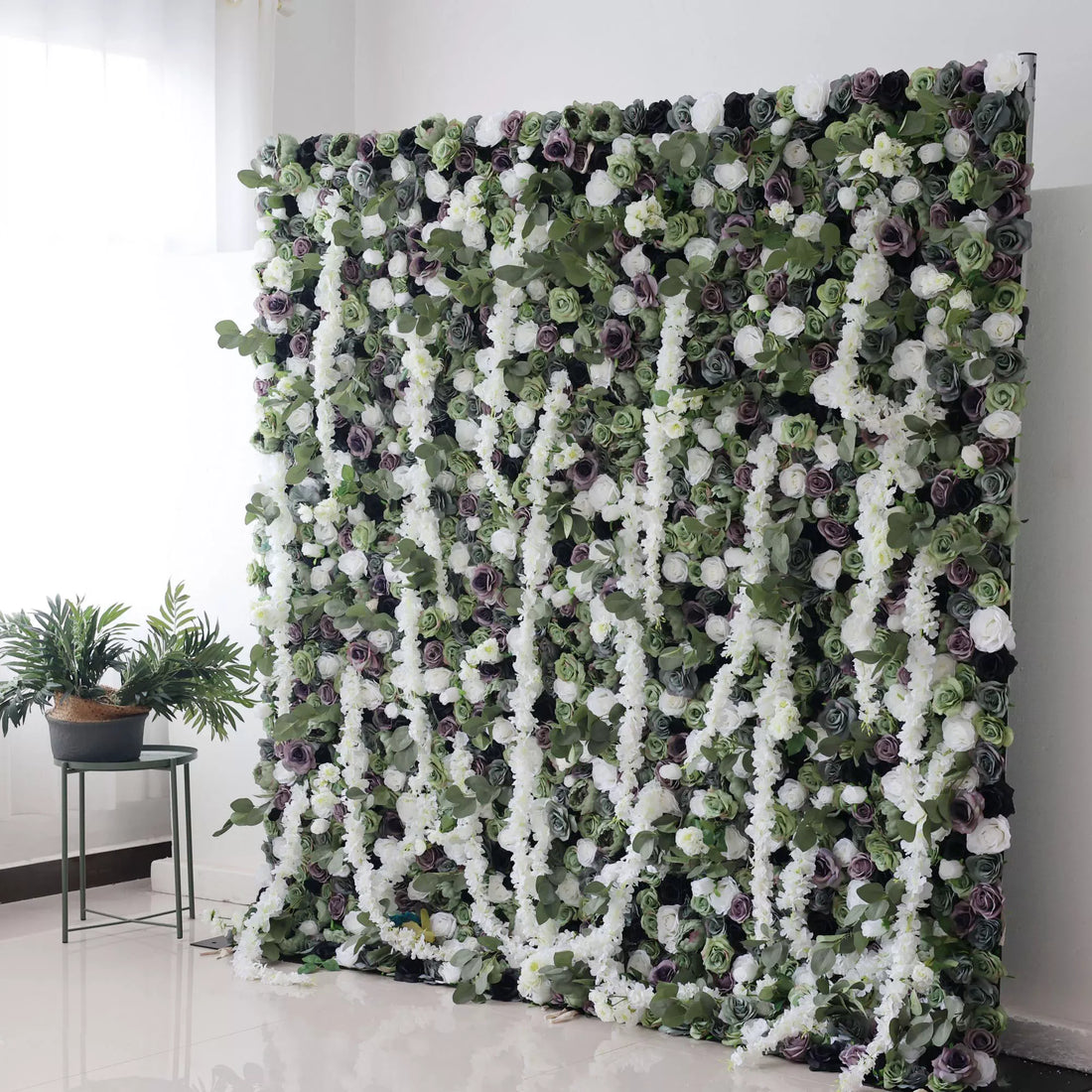 This is a large artificial flower wall with a lush, green - based color scheme. It features white flowers, purple roses, and green foliage, creating a dense, natural - looking display. Paired with potted plants on the side, it adds an elegant, verdant touch, perfect for decorative event backdrops.