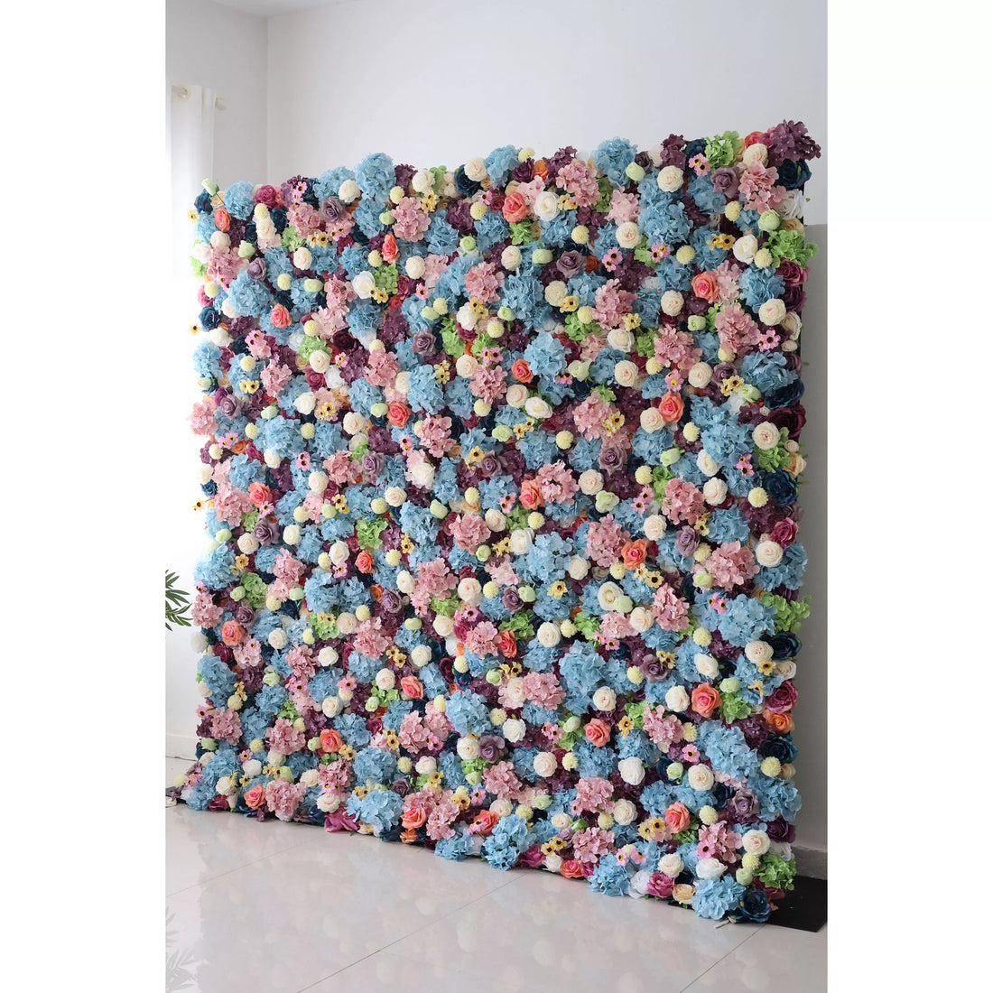 This is a large, vibrant artificial flower wall. It features a dense mix of blue hydrangeas, white roses, pink blossoms, and small yellow flowers, interspersed with green foliage. The colorful, lush arrangement creates a lively, eye - catching display, perfect for event backdrops, adding a cheerful and decorative touch.