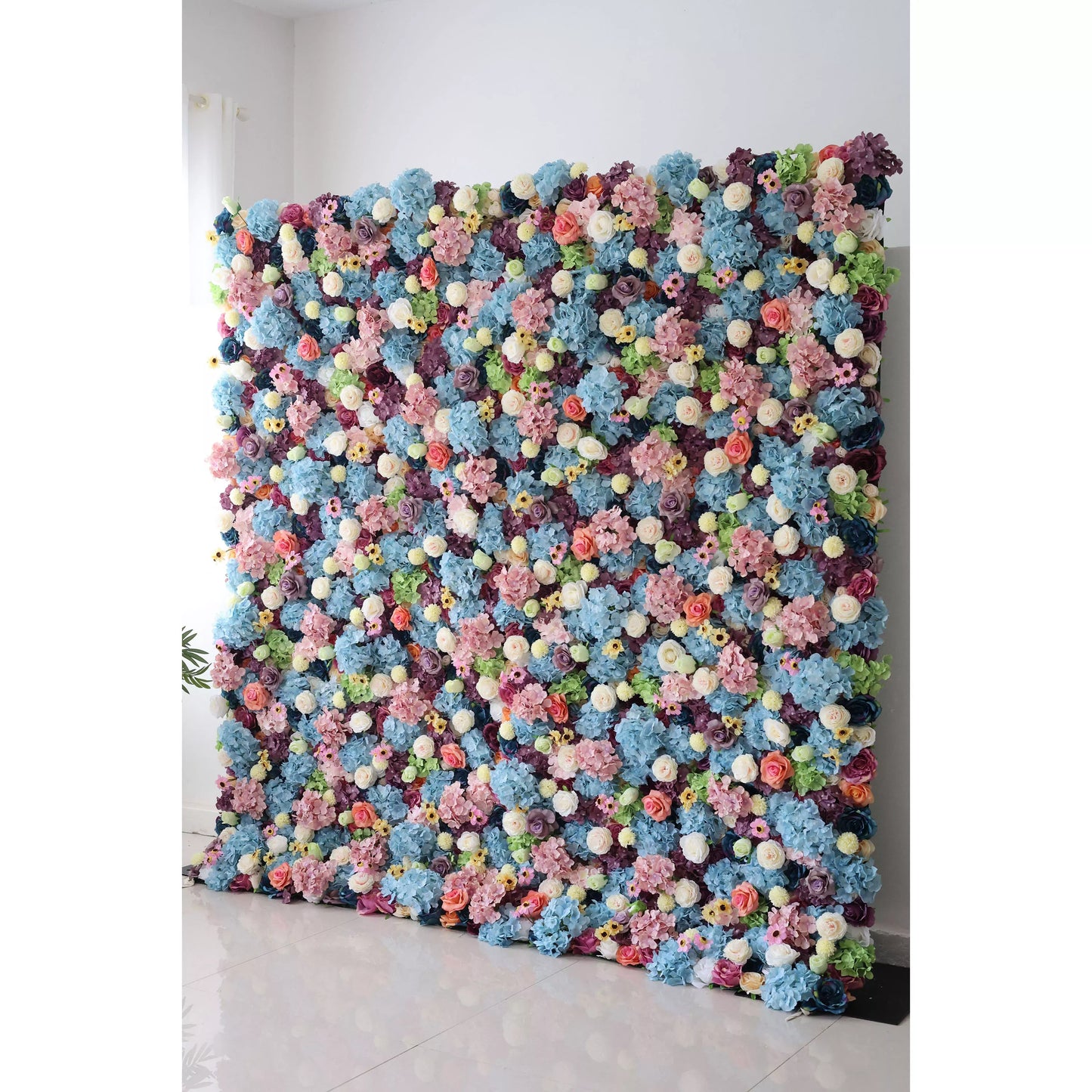 This is a large, vibrant artificial flower wall. It features a dense mix of blue hydrangeas, white roses, pink blossoms, and small yellow flowers, interspersed with green foliage. The colorful, lush arrangement creates a lively, eye - catching display, perfect for event backdrops, adding a cheerful and decorative touch. 