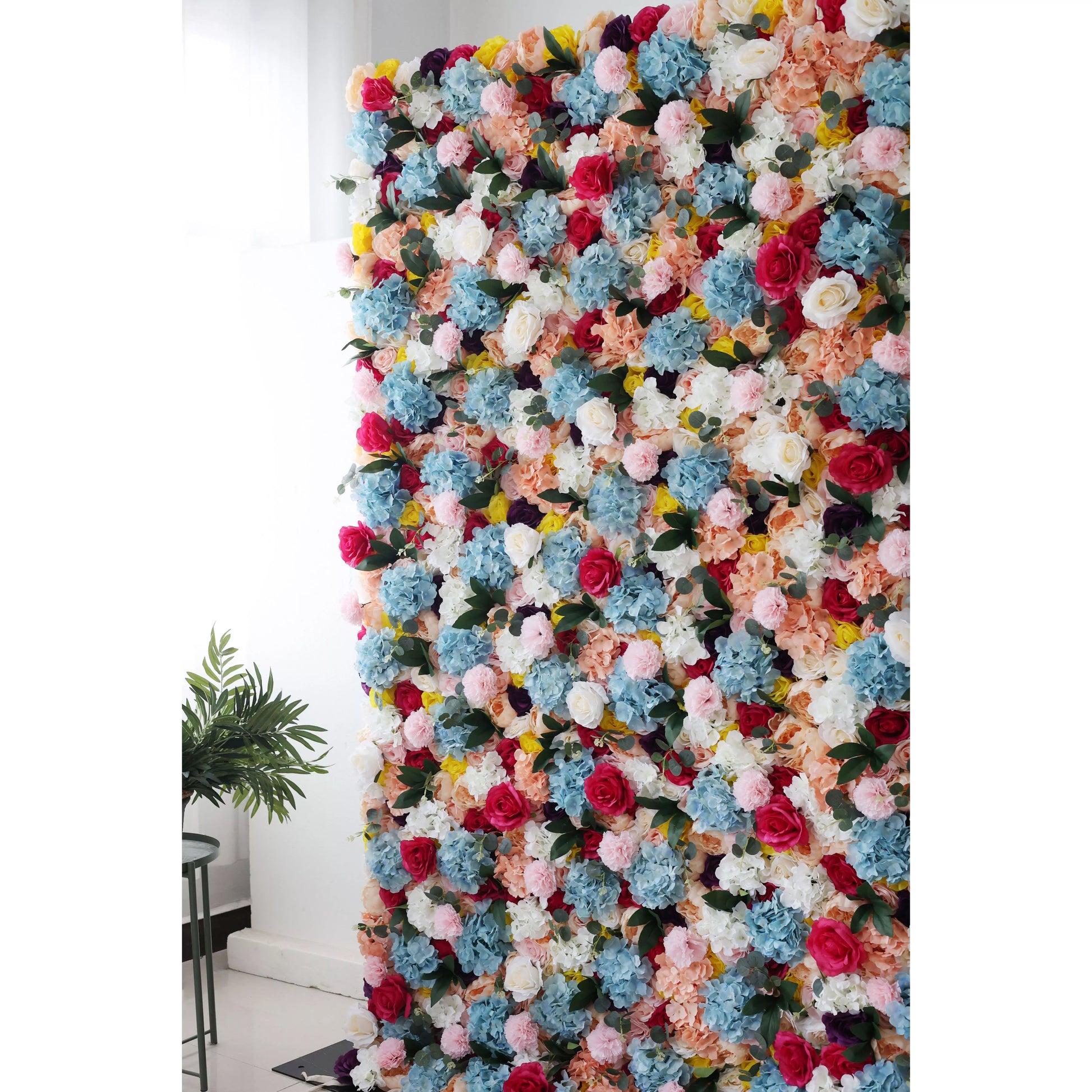 This is a large, vibrant artificial flower wall. It features a dense mix of colorful blooms—blue hydrangeas, pink roses, white flowers, and yellow blossoms. The lush arrangement creates a lively, eye - catching display, perfect for event backdrops, adding a cheerful and decorative touch to the space. 