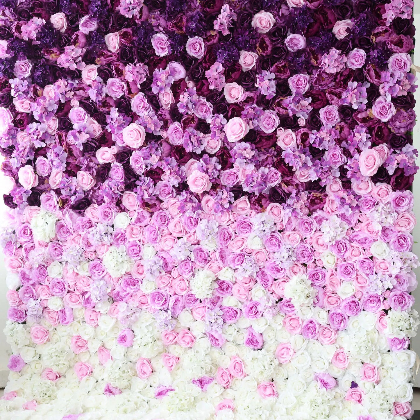 This flower wall is a stunning arrangement of artificial flowers, showcasing a gradient of colors from deep purple at the top, transitioning through various shades of pink, to white at the bottom. Comprising roses and other delicate blooms, it creates a lush, romantic texture. Ideal for weddings, parties, or other special events, it serves as an enchanting backdrop, exuding elegance and charm. 