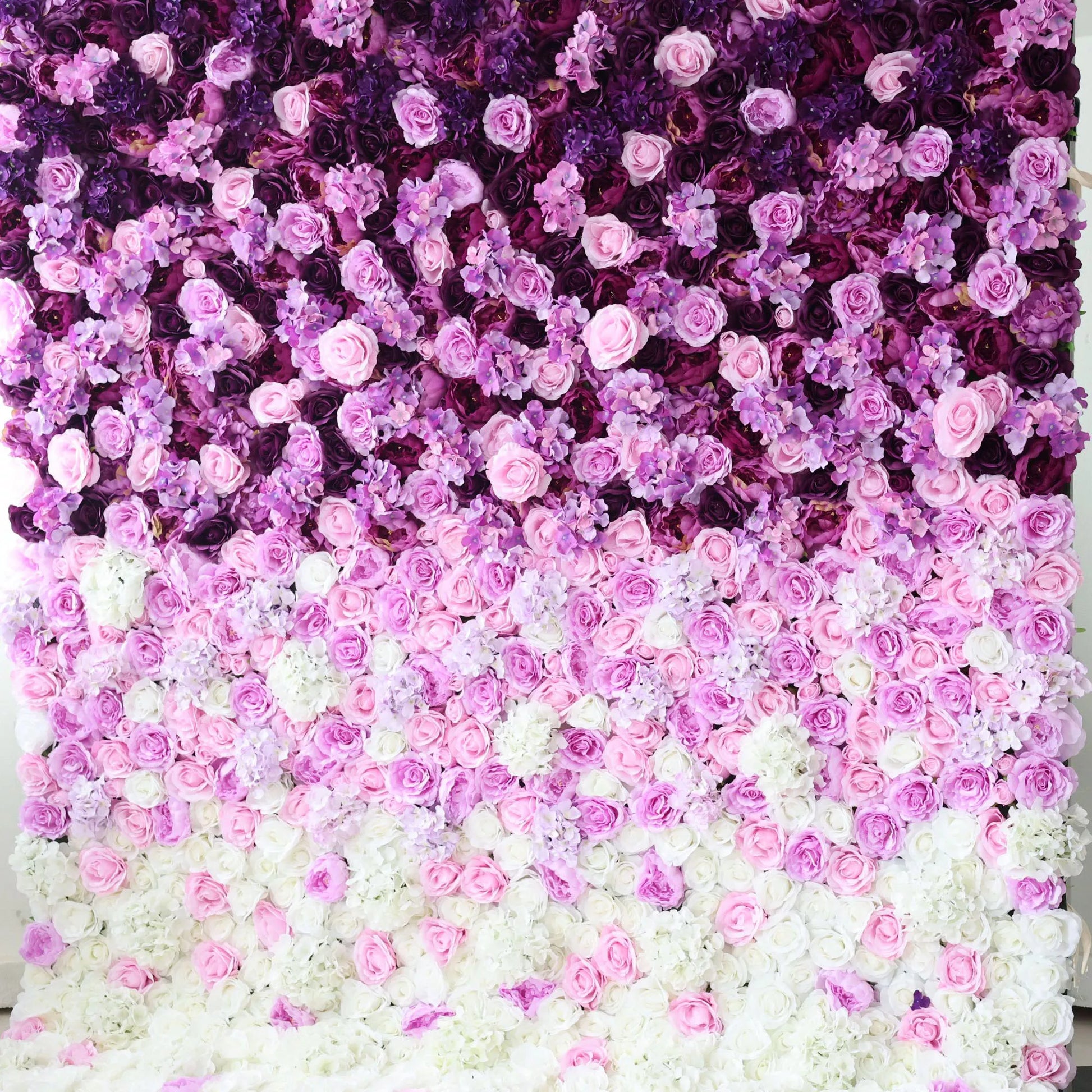 This flower wall is a stunning arrangement of artificial flowers, showcasing a gradient of colors from deep purple at the top, transitioning through various shades of pink, to white at the bottom. Comprising roses and other delicate blooms, it creates a lush, romantic texture. Ideal for weddings, parties, or other special events, it serves as an enchanting backdrop, exuding elegance and charm. 