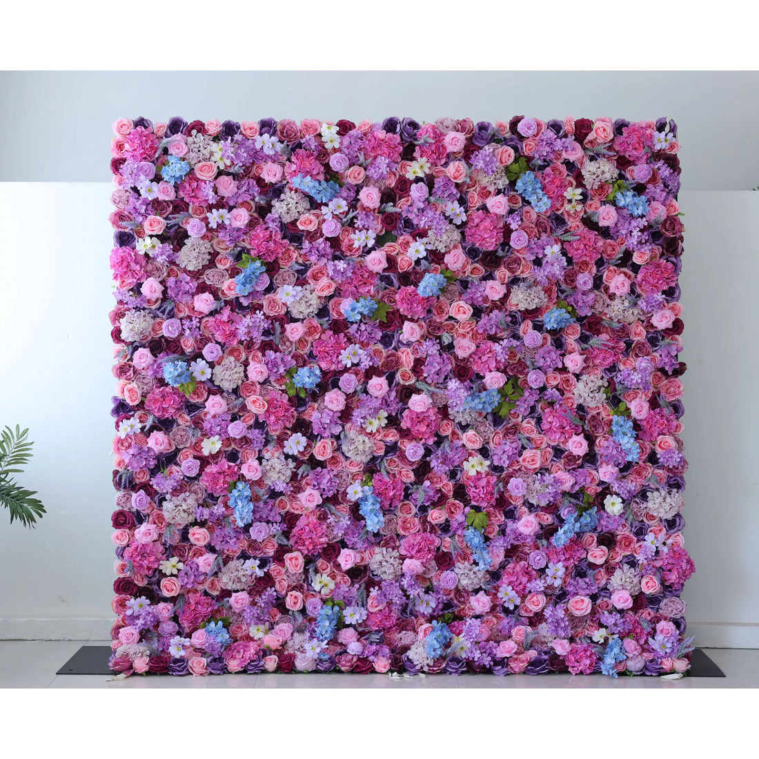 This flower wall is a vibrant, dense tapestry of artificial blooms. A rich mix of pink, purple, lavender, and blue flowers—roses, hydrangeas, and delicate wildflowers—creates a lush, romantic texture. The harmonious color palette and varied floral shapes make it perfect for weddings or events, exuding charm and elegance.