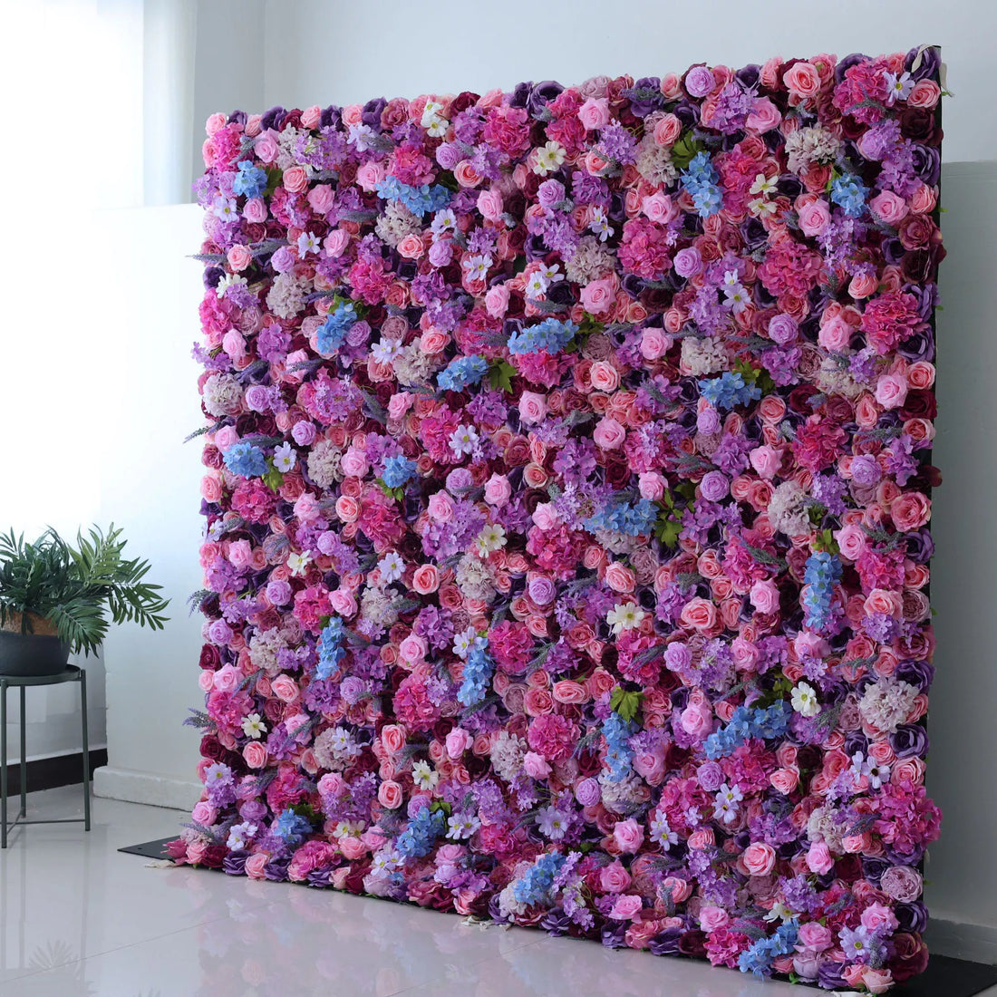 This flower wall is a vibrant, dense display of artificial blooms. A harmonious blend of pink, purple, lavender, and blue flowers, including roses and hydrangeas, creates a dreamy, colorful tapestry. The lush arrangement and varied textures make it perfect for weddings or events, exuding romance and charm.