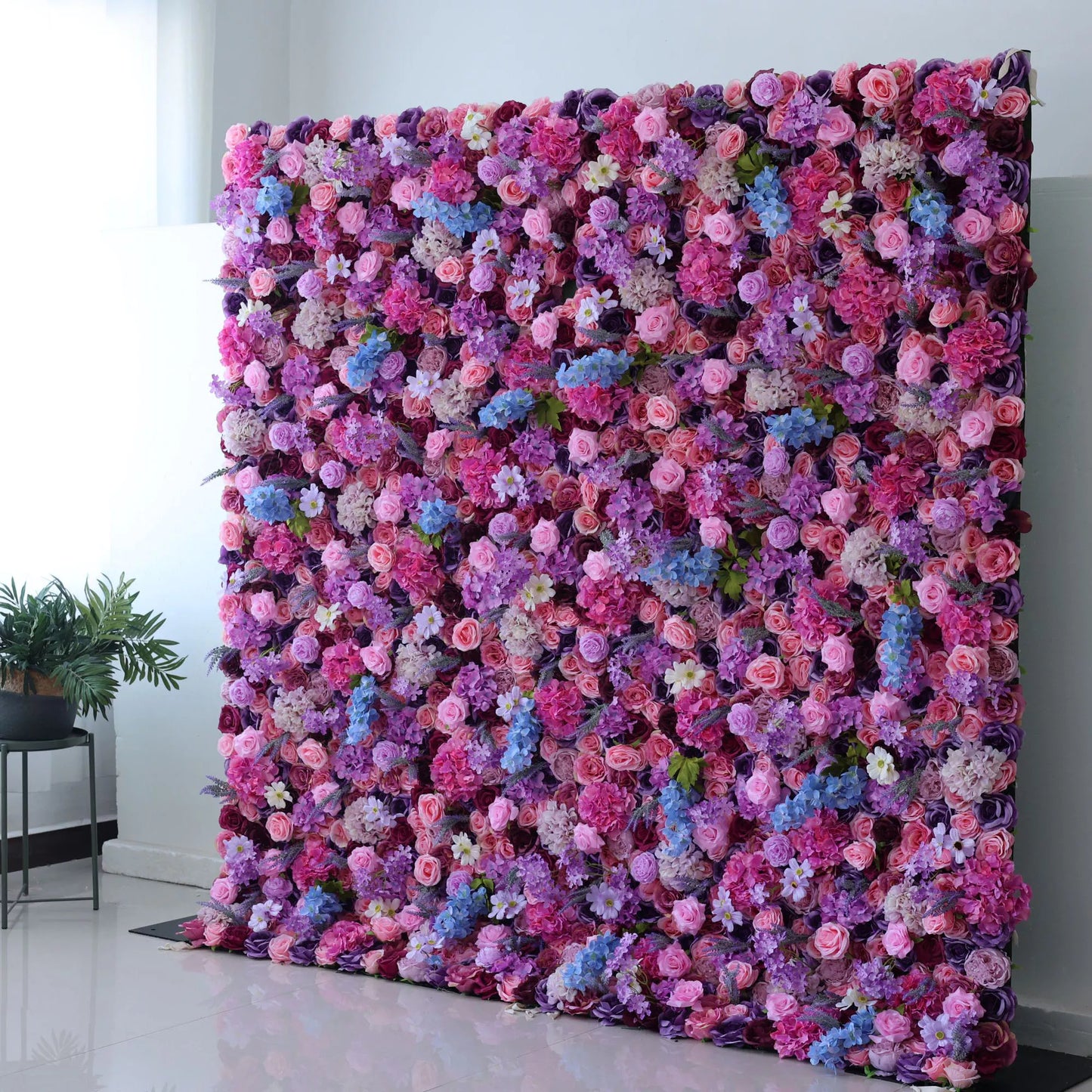 This flower wall is a vibrant, dense display of artificial blooms. A harmonious blend of pink, purple, lavender, and blue flowers, including roses and hydrangeas, creates a dreamy, colorful tapestry. The lush arrangement and varied textures make it perfect for weddings or events, exuding romance and charm. 