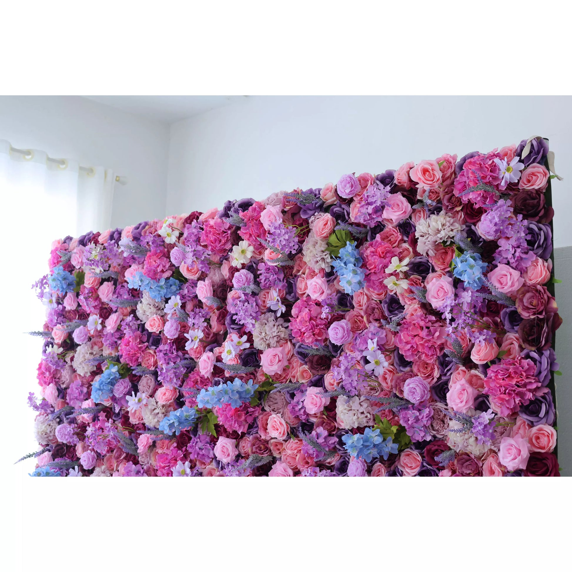 This is a lush artificial flower wall. It features a vibrant mix of pink, purple, lavender, and blue blooms—roses, hydrangeas, and small white flowers. The dense arrangement creates a romantic, colorful backdrop, perfect for events like weddings, adding an elegant and charming atmosphere. 