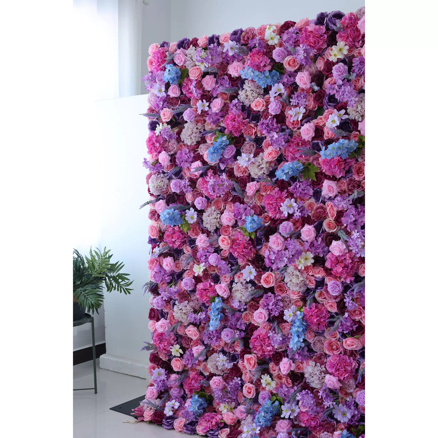 This is a tall, lush artificial flower wall. It features a vibrant mix of pink, purple, lavender, and blue blooms, including roses, hydrangeas, and small white flowers. The dense arrangement creates a romantic, colorful backdrop, complemented by a green plant on the side. Ideal for decorative event settings. 