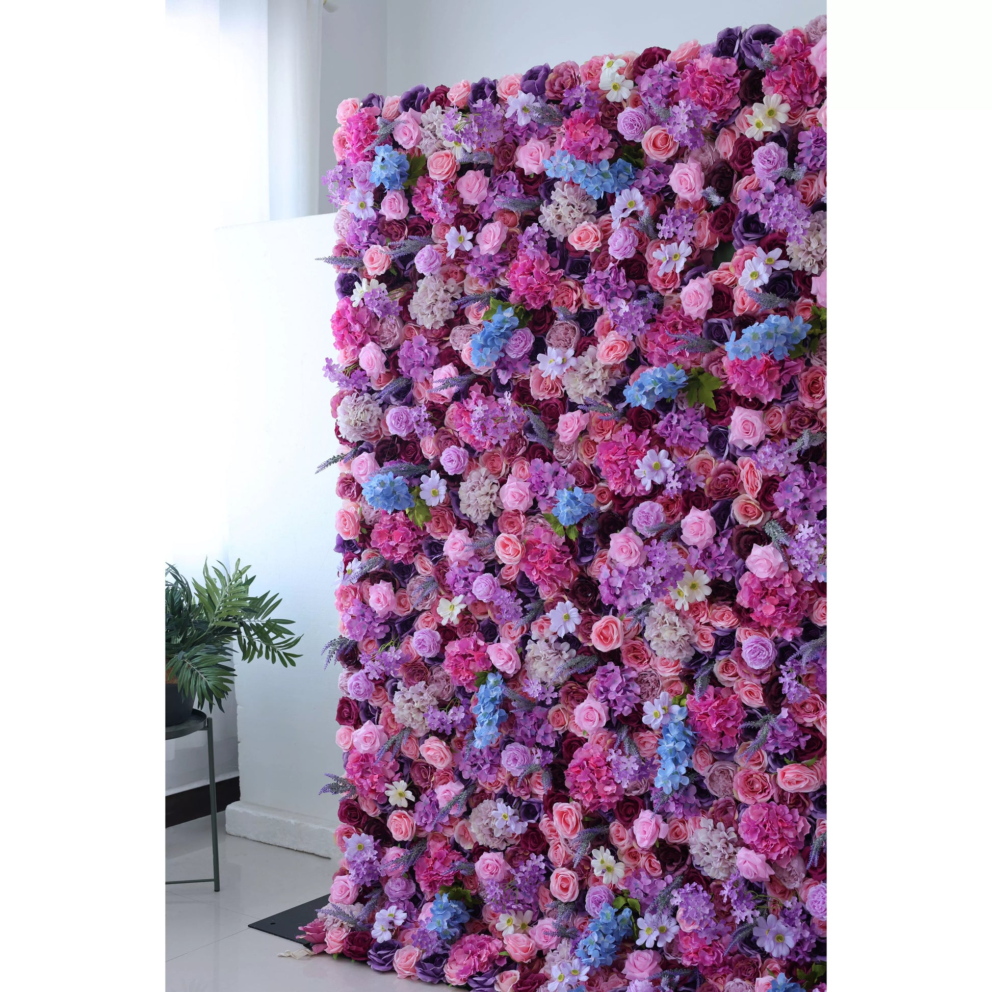 This is a tall, lush artificial flower wall. It features a vibrant mix of pink, purple, lavender, and blue blooms, including roses, hydrangeas, and small white flowers. The dense arrangement creates a romantic, colorful backdrop, complemented by a green plant on the side. Ideal for decorative event settings. 