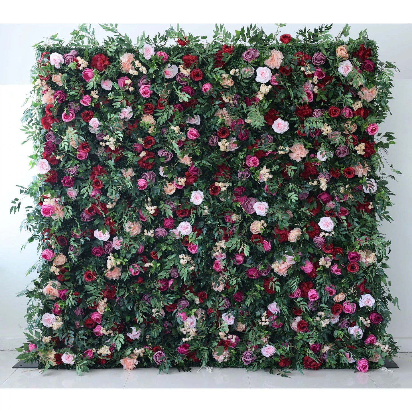 A large artificial flower wall showcases a vibrant mix of roses in pink, red, purple, and peach, interspersed with green foliage and small white blooms. It creates a lush, romantic backdrop, ideal for events or decorative spaces, exuding natural beauty and elegance. 