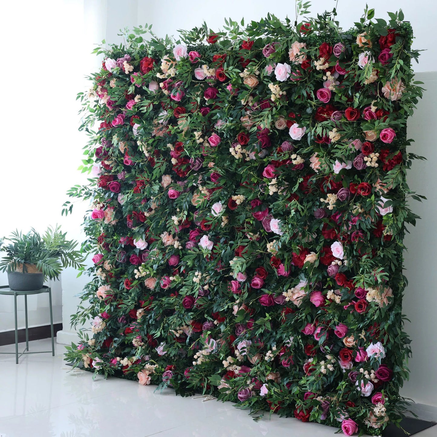 A large artificial flower wall bursts with vibrant blooms—pink, red, purple roses, plus small white flowers—all nestled in lush green foliage. It creates a lush, romantic backdrop, perfect for events or decorative spaces, adding a touch of natural beauty indoors. 