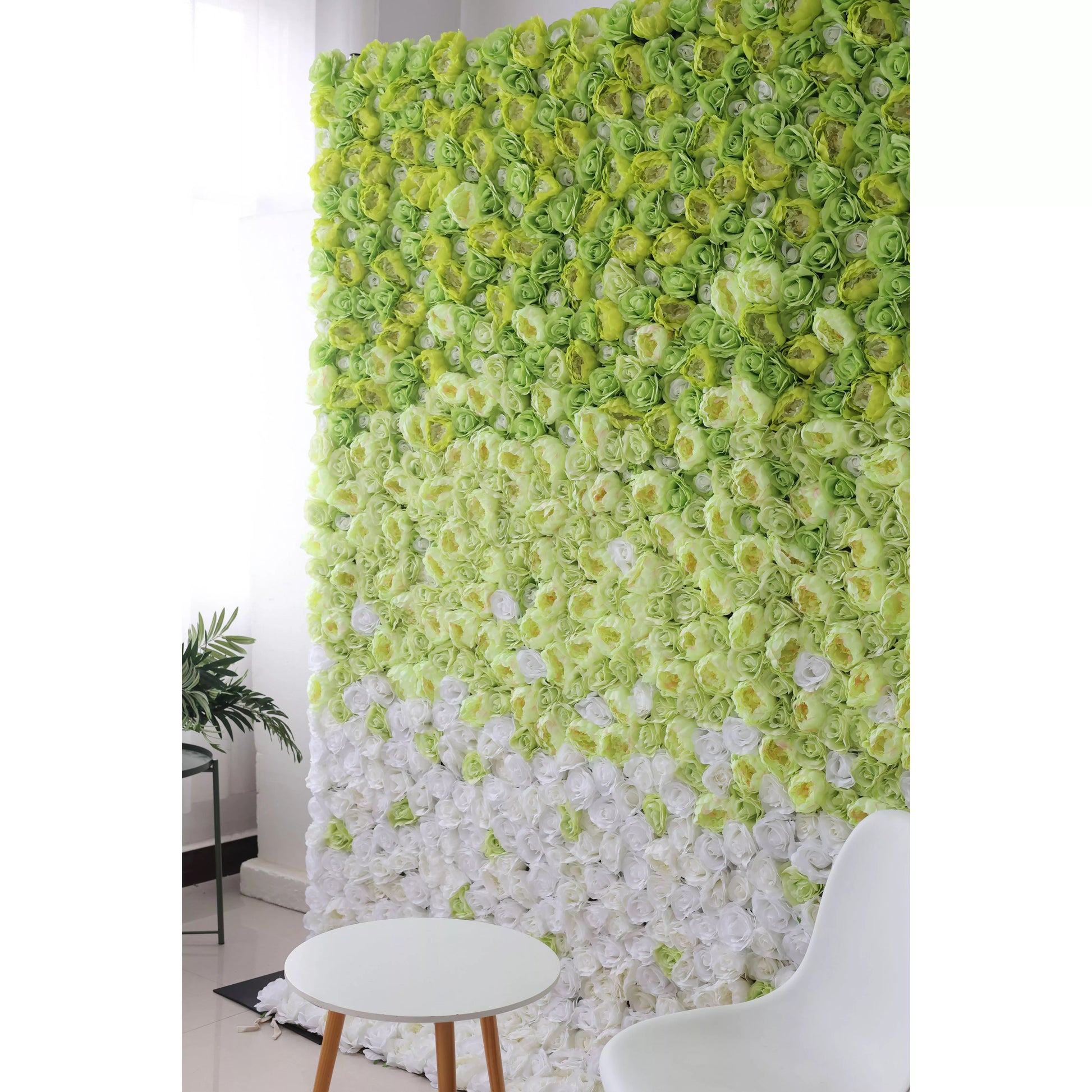 This flower wall showcases a stunning gradient of artificial roses, transitioning from vibrant green at the top to pure white at the bottom. The dense arrangement creates a lush, textured backdrop. Complemented by a minimalist white table and chair, it exudes a fresh, elegant charm, perfect for photo ops or event decor. 