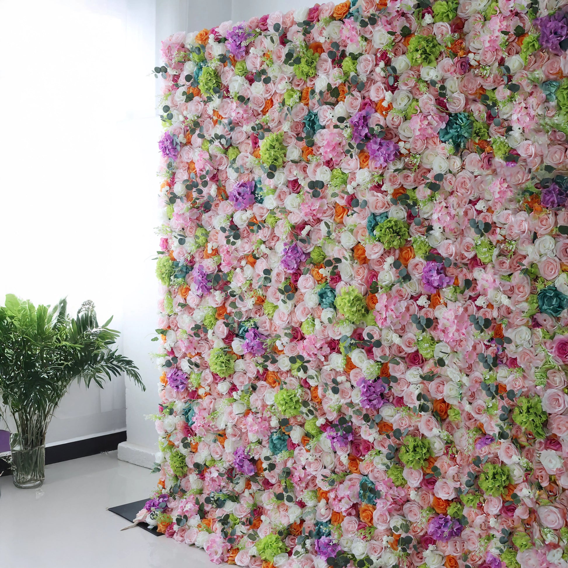 This is a vibrant, multi-colored artificial flower wall backdrop from **Valar Flowers**, densely packed with a colorful lush mix of blooms in soft pink, white, orange, purple, and turquoise, complemented by green hydrangeas and eucalyptus leaves. Set indoors against a white wall, it creates a lively, romantic, and eye-catching aesthetic ideal for wedding decor, photo backdrops, or special event styling.
