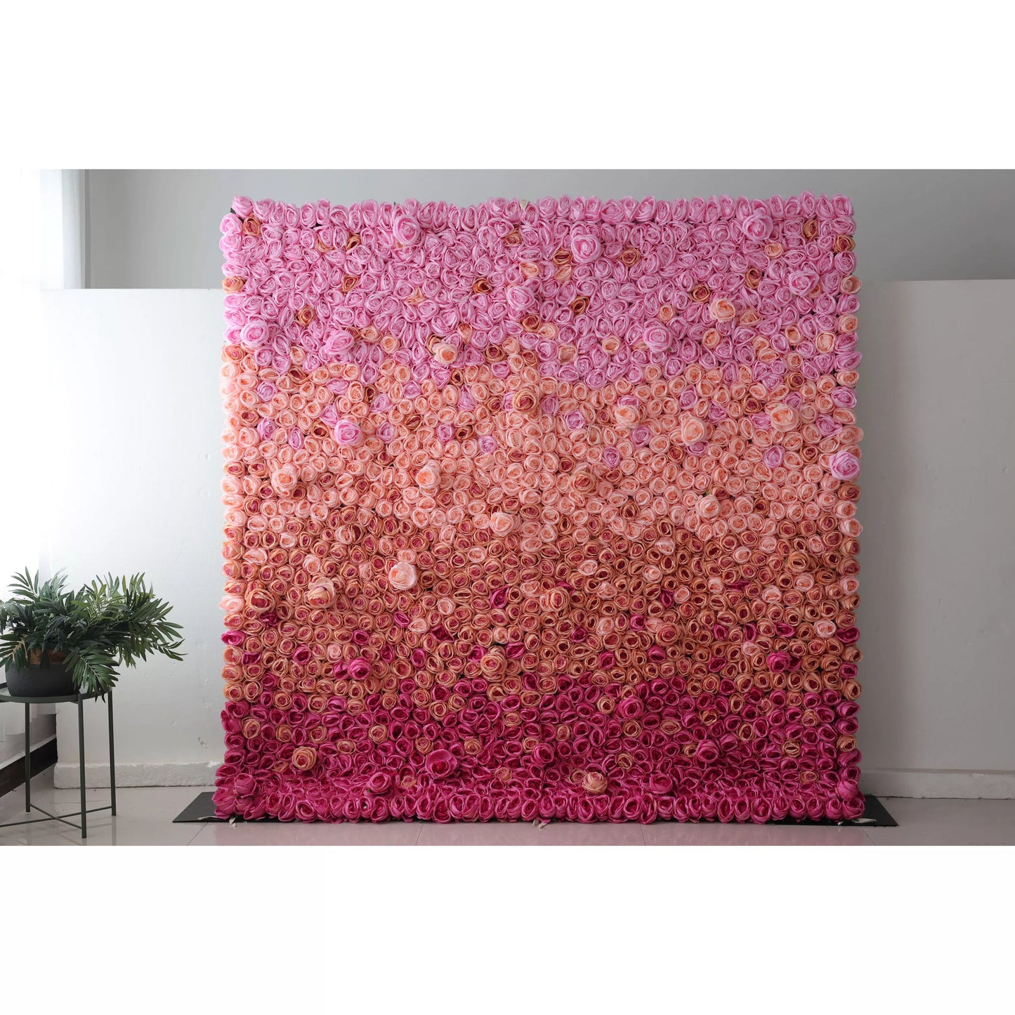 This picture showcases a large artificial flower wall, a beautiful gradient of pink shades. It's densely packed with rose - like blooms, transitioning from soft pink at the top to deeper pink at the bottom. The uniform, lush arrangement creates a romantic, eye - catching display, perfect for event backdrops. A small plant on the side complements the setup, adding a touch of natural greenery to the scene. 