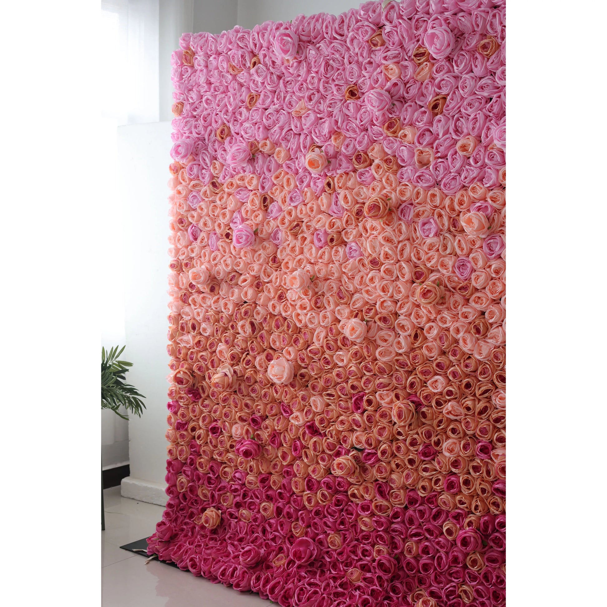 This picture features a large artificial flower wall, a beautiful gradient of pink tones. It's densely packed with rose - like blooms, transitioning from soft pink at the top to deeper pink at the bottom. The uniform, lush arrangement creates a romantic and eye - catching display, perfect for event backdrops. A small green plant on the side adds a touch of natural contrast, enhancing the overall decorative appeal. 