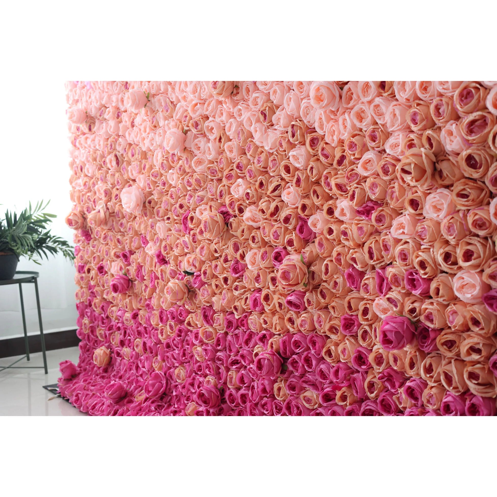 This picture presents a large artificial flower wall, a stunning gradient of pink tones. It’s densely packed with rose - like blooms, transitioning from soft peach - pink at the top to deeper pink at the bottom. The uniform, lush arrangement creates a romantic, eye - catching display, perfect for event backdrops. A small green plant on a side table adds a touch of natural contrast, enhancing the overall decorative appeal of the space. 