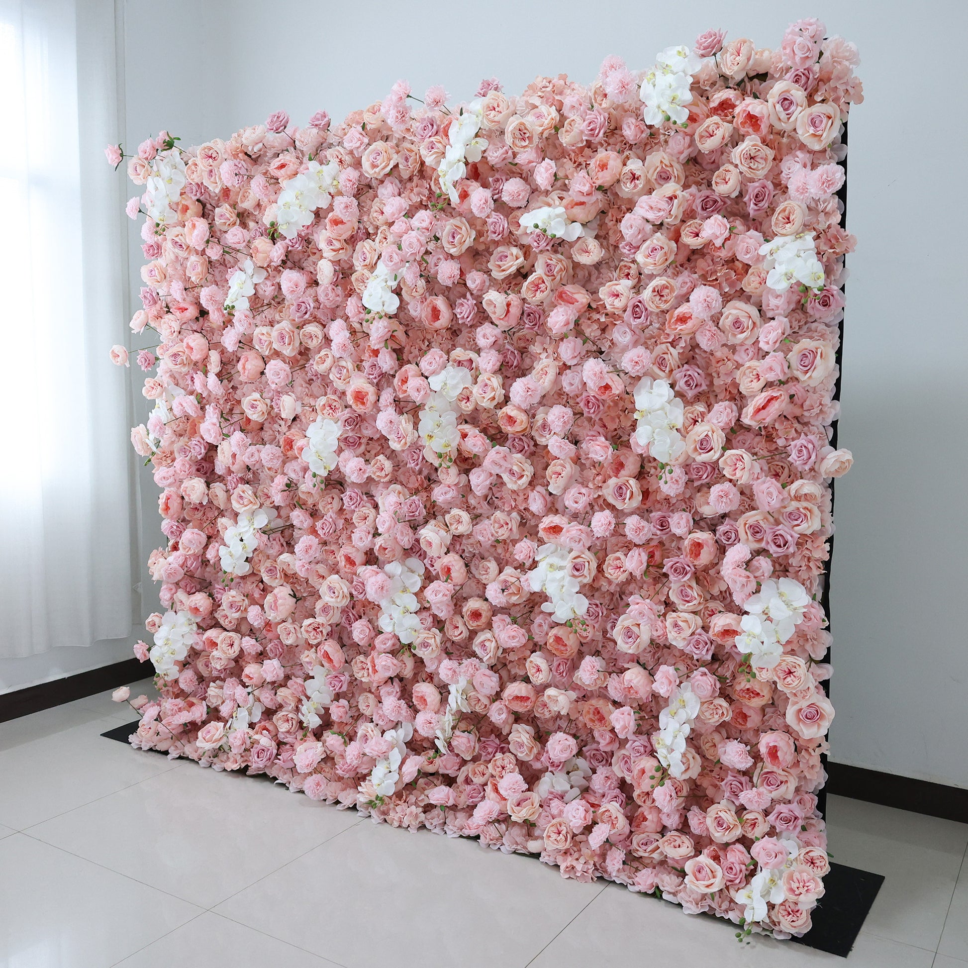 This image showcases a beautiful floral wall by Valar Flowers, set indoors against a plain white wall and tiled floor. It features a dense arrangement of artificial pink and white flowers, creating a soft and romantic display. The backdrop is perfect for events, with the delicate hues and lush texture adding a touch of elegance, ideal for enhancing the ambiance of weddings or photo shoots. 