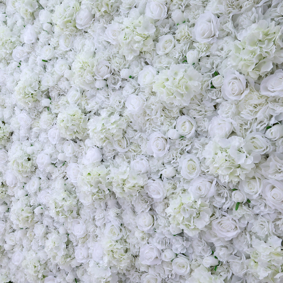 A dense arrangement of artificial white flowers forms a lush wall. It features various blooms like roses and hydrangeas, with subtle green leaves peeking through. The pure white hues create an elegant, romantic look, perfect for events such as weddings, bridal showers, or upscale parties needing a sophisticated backdrop.