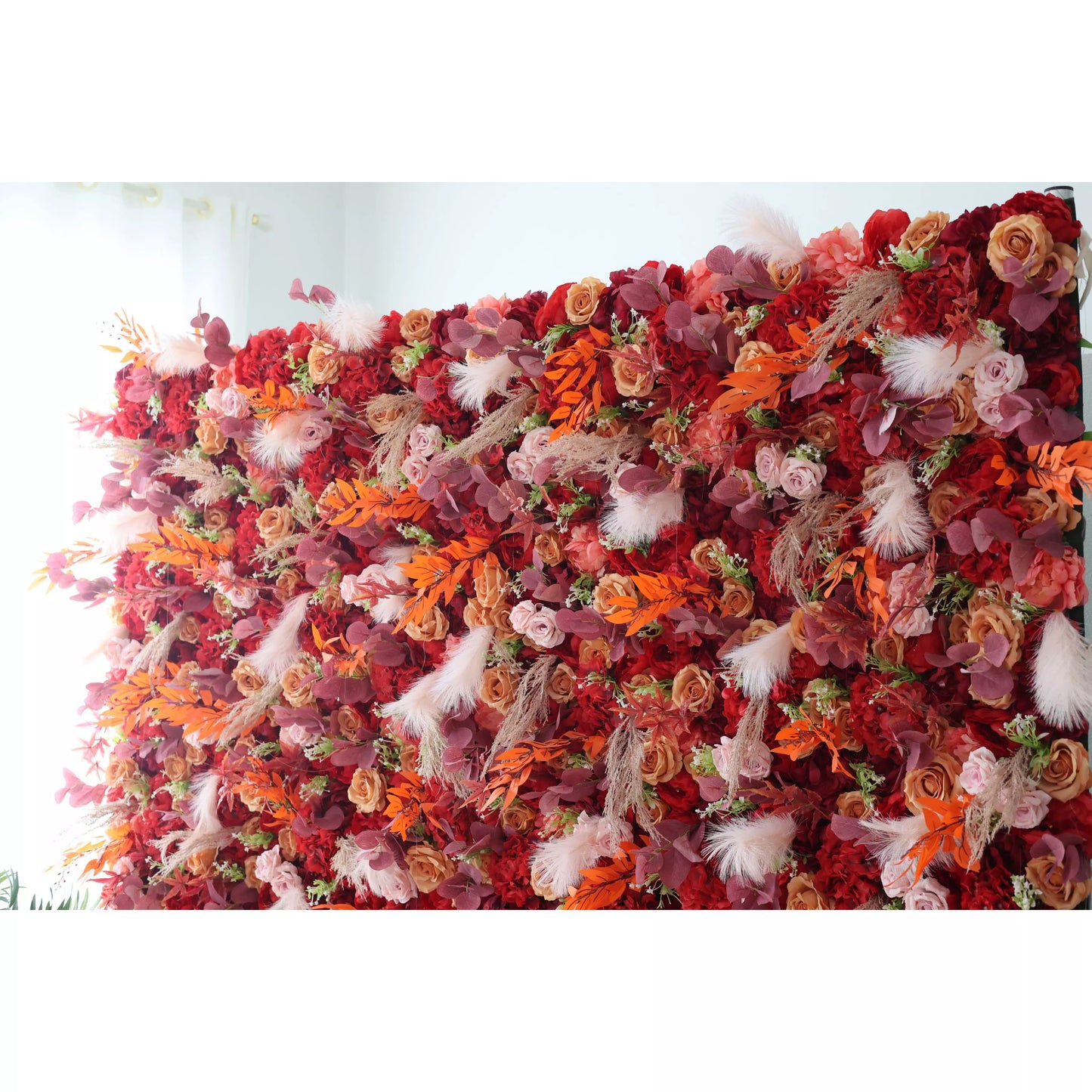 This flower wall is a rich and vibrant display, densely filled with artificial flowers. It features a warm color palette, combining deep reds, oranges, and soft pinks. Interspersed among the blooms are decorative elements like white feathers and various foliage, adding texture and visual interest. Ideal for weddings or events, it exudes a passionate and elegant atmosphere, serving as a stunning backdrop. 